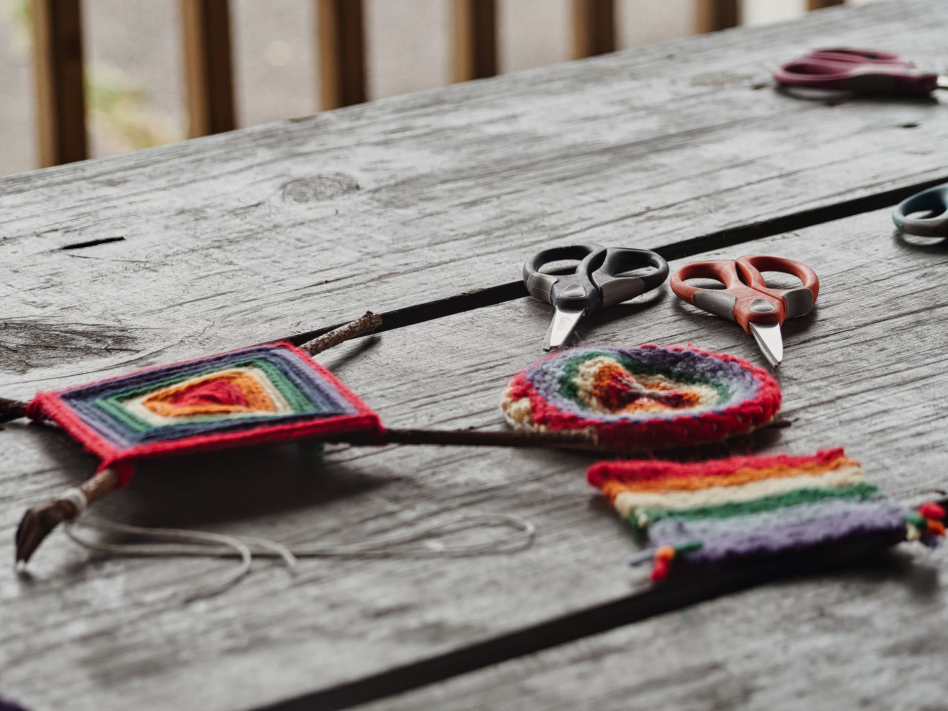 Small woven yarn pieces in rainbow striped and concentric diamond patterns laid across a weathered grey wooden table, alongside two pairs of scissors — one grey-handled, one red — and a thin stick running diagonally through the frame. A third pair of scissors and a spool sit blurred in the upper right background.