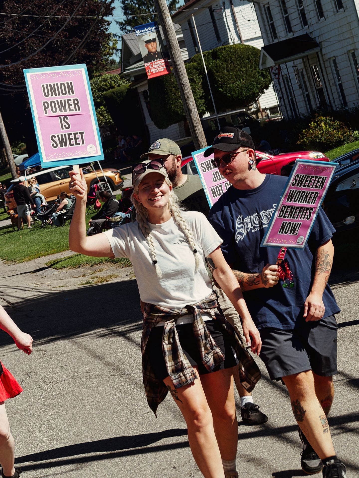 Three people marching down a sunny residential street, each holding pink-and-teal signs reading "UNION POWER IS SWEET" and "SWEETEN WORKER BENEFITS NOW!" — a smiling woman in the foreground with long blonde braids and a flannel tied at the waist raises her sign overhead, while two others in baseball caps and navy tees walk behind her.
