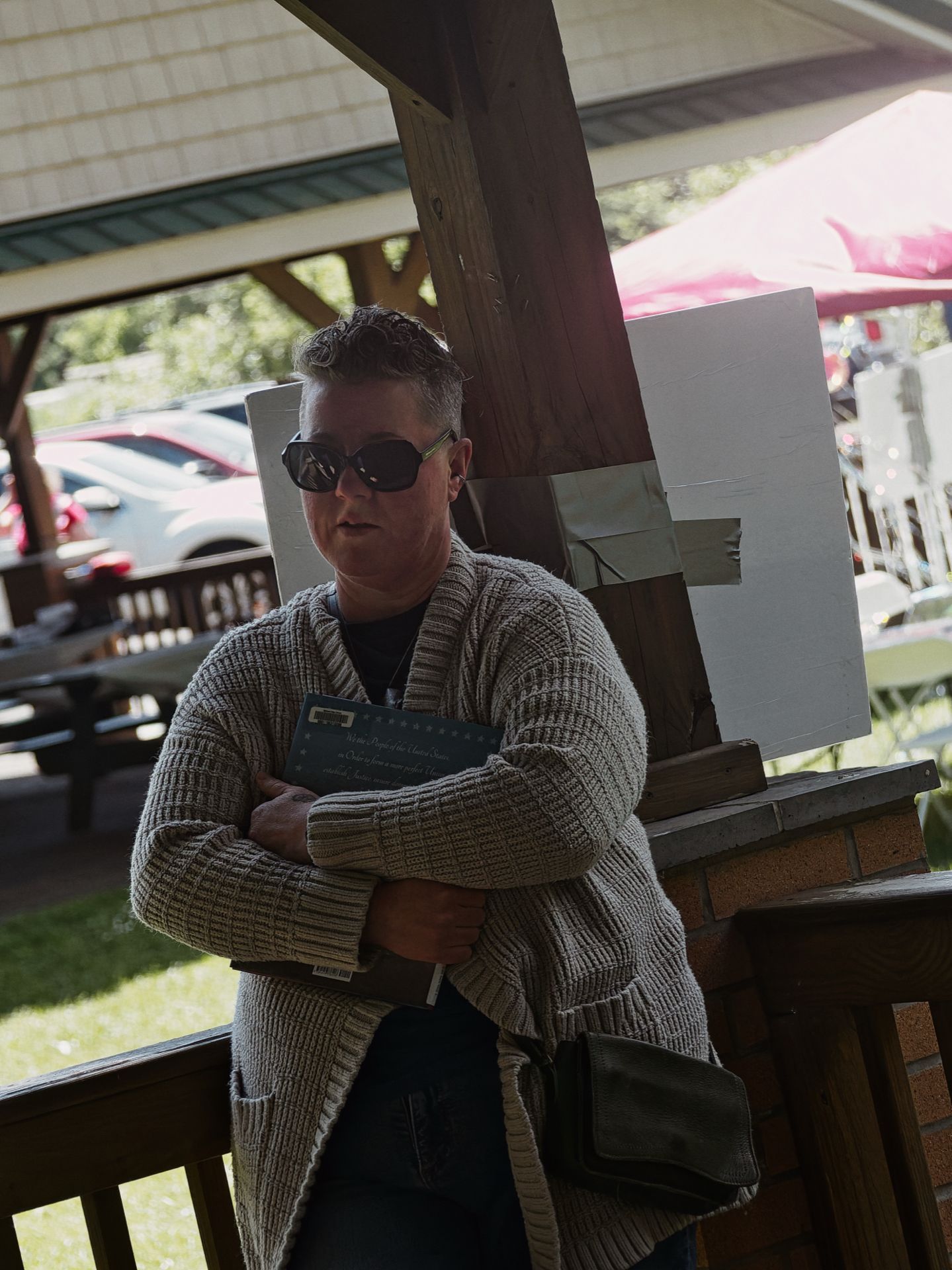 Person with short gray hair and dark oversized sunglasses, standing arms-crossed beneath a wooden pavilion structure, clutching a dark navy booklet against their chest while wearing a chunky knit gray cardigan. A white foam board taped to a wooden easel stands behind them to the right, with parked cars and green trees blurred in the background to the left.