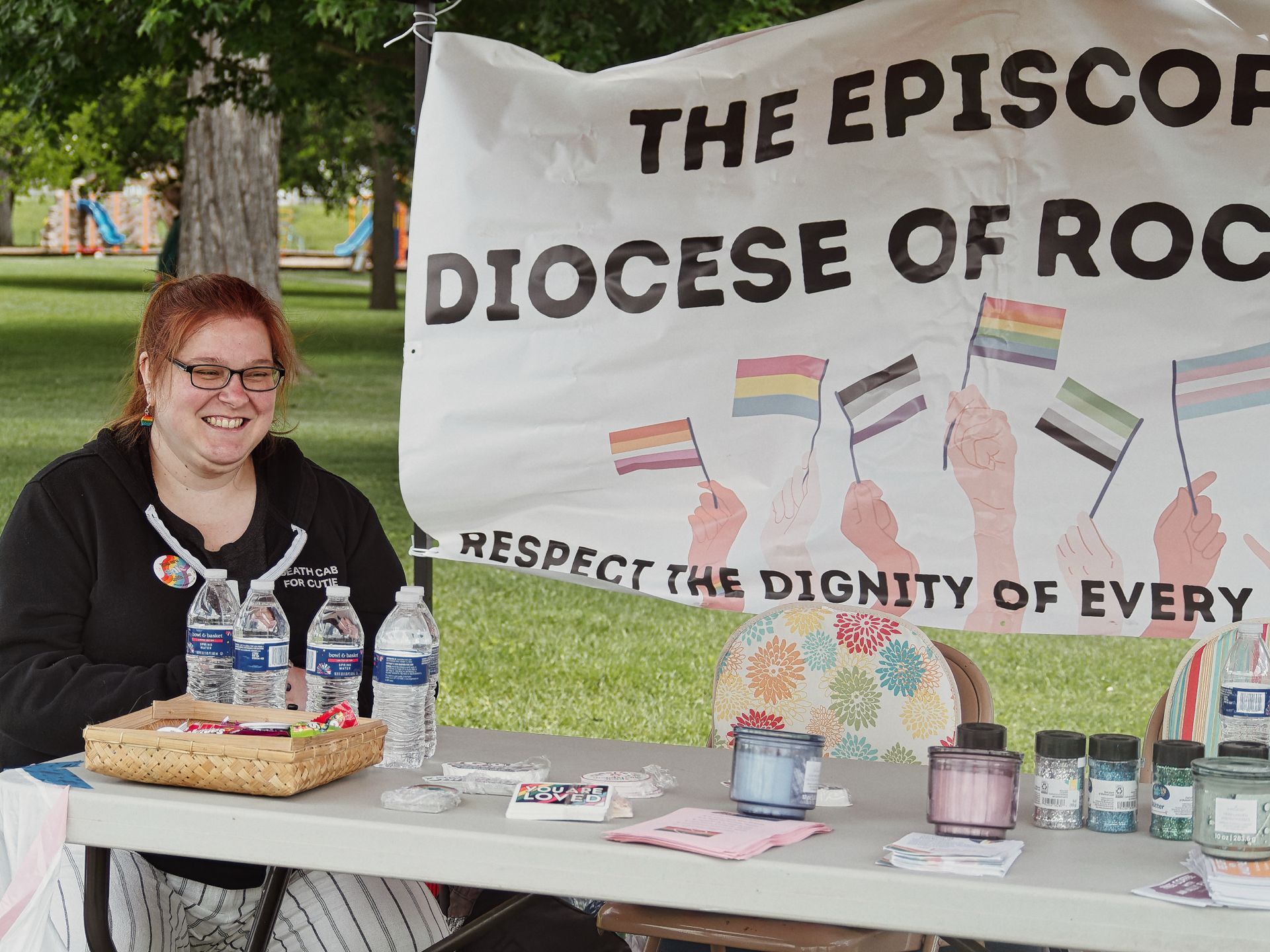 A smiling woman with red hair and glasses, wearing a black Death Cab for Cutie hoodie, sits at an outdoor tabling booth in a park. The table in front of her holds water bottles, a wicker tray of candy, glitter jars, and a small card reading "YOU ARE LOVED," with a partially visible banner behind her reading "THE EPISCOPAL DIOCESE OF ROC — RESPECT THE DIGNITY OF EVERY," illustrated with hands holding various Pride flags.