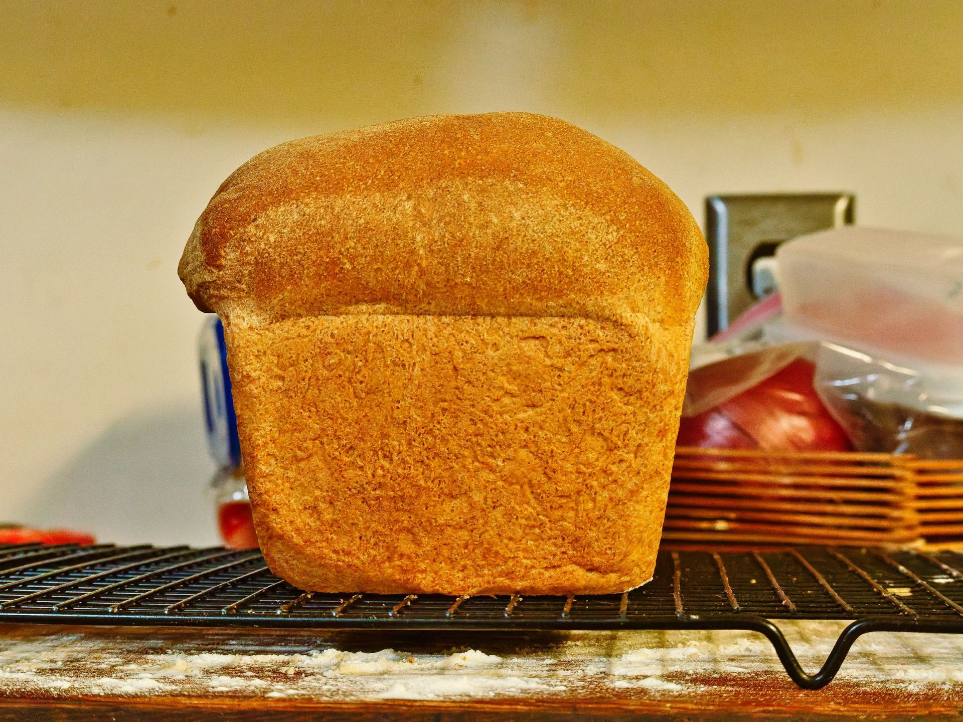 Golden-brown homemade sandwich loaf with a domed, evenly baked crust sitting on a black wire cooling rack dusted with flour, against a cream-colored wall. A basket of red onions in a plastic bag and a blue bag sit blurred in the shallow depth of the background.