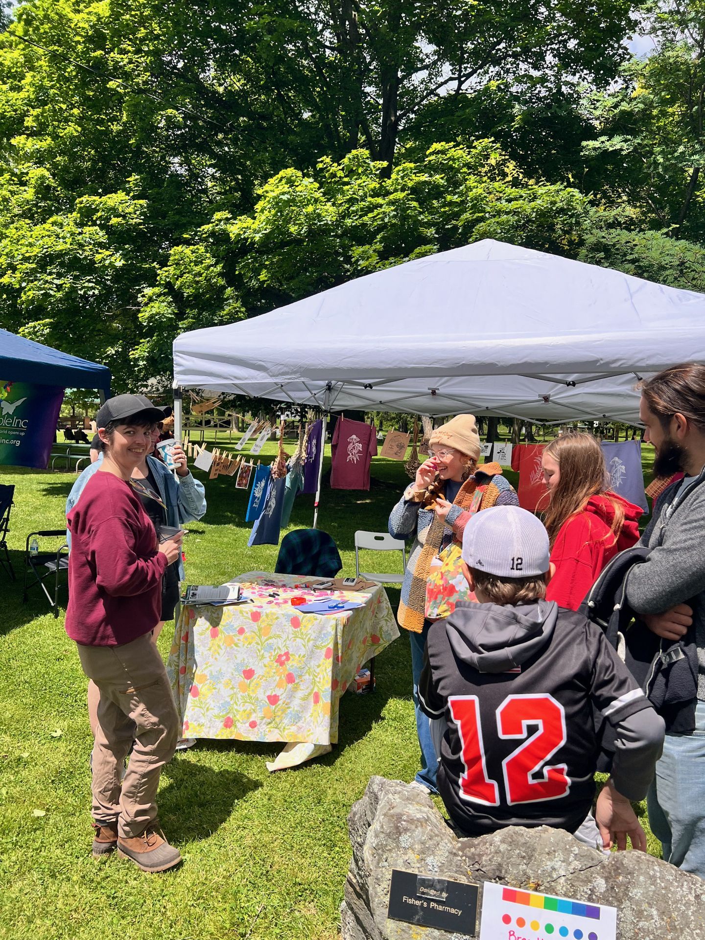 Outdoor craft fair booth beneath a white pop-up canopy on a sunny green lawn, with colorful printed fabric panels and cards strung along a line in the background. A vendor in a maroon cardigan and black cap smiles at left, while a small group of attendees — including a child in a "12" jersey — clusters around a floral-tableclothed table at center; a sign reading "Fisher's Pharmacy" with a rainbow color strip sits in the foreground on a stone.