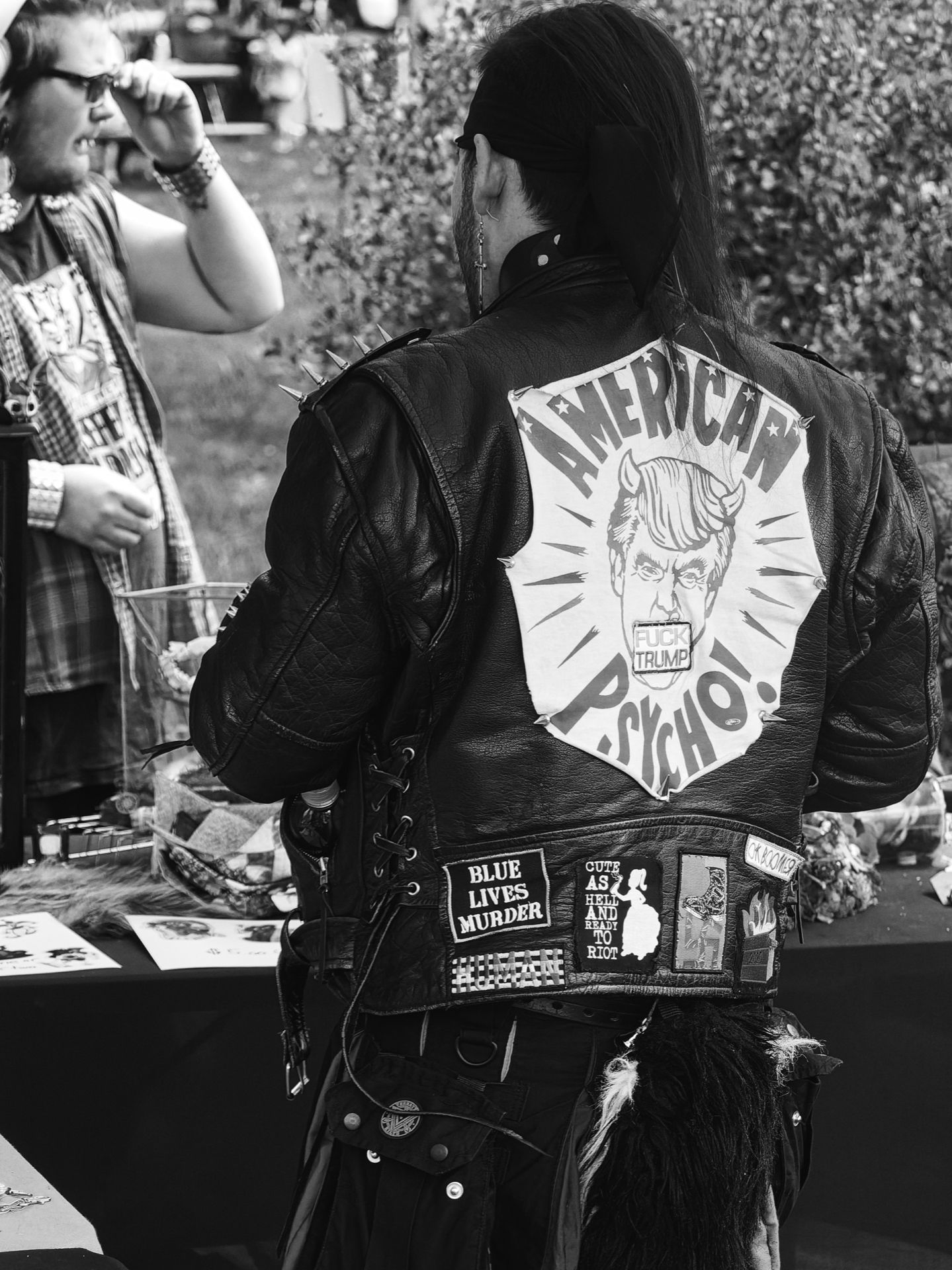 Black-and-white vertical shot of a person seen from behind, wearing a spiked black leather jacket with a large hand-painted back patch depicting a devil-horned caricature of Donald Trump with the text "AMERICAN PSYCHO!" and "FUCK TRUMP." Several smaller patches line the lower back, reading "BLUE LIVES MURDER," "CUTE AS HELL AND READY TO RIOT," and others partially visible. In the background to the left, another person holds a microphone to their mouth near what appears to be a vendor table, with dense foliage blurred behind them.