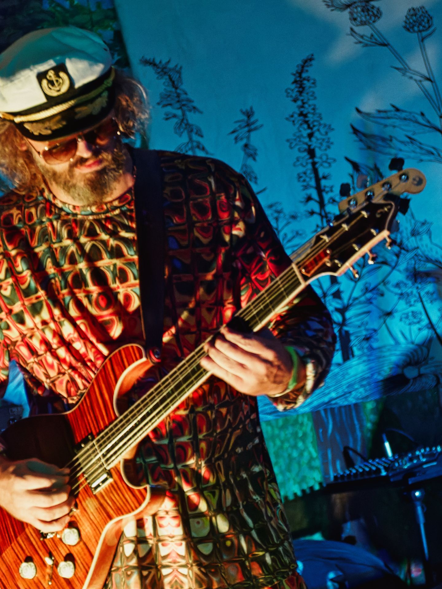 Vertical shot of a bearded guitarist wearing a white captain's hat with an anchor emblem and tinted sunglasses, playing a warm-toned hollow-body electric guitar under dramatic amber stage lighting, his psychedelic red-and-green patterned suit popping against a cool blue backdrop printed with silhouetted wildflowers and botanical shapes.