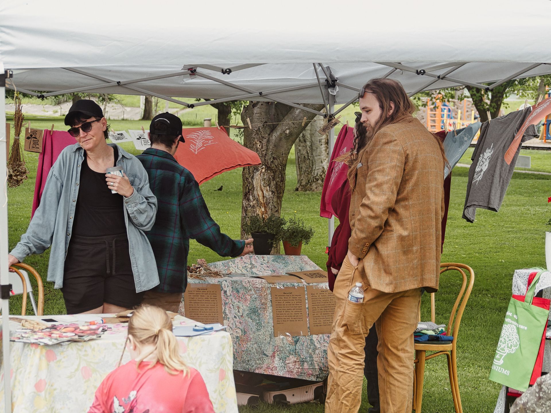 Outdoor craft market booth under a white canopy tent, with printed and dyed garments — burgundy and orange-red — hanging on a clothesline strung between trees in the background. On the left, a person in a denim jacket and black cap holds a small cup; center, someone in a green flannel leans over a floral-cloth-covered table; right, a long-haired man in a tan tweed blazer and matching pants stands with hands in pockets, a water bottle tucked at his side. In the foreground, a blonde person in a pink top sits at a separate table covered in small items, with a green tote bag reading "NIUMS" visible at the far right.