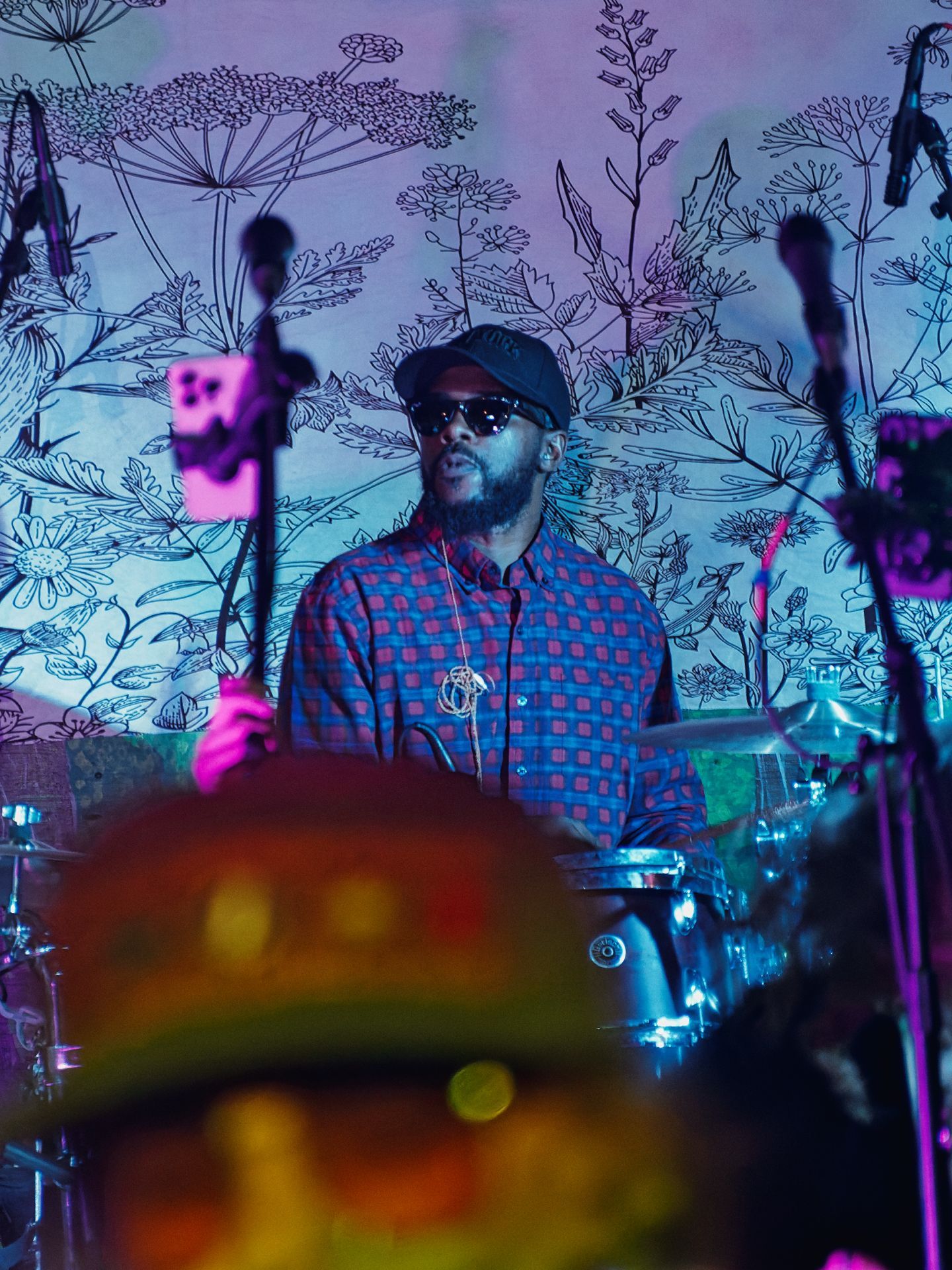 Vertical shot of a drummer mid-performance, wearing a dark cap, sunglasses, and a red-and-blue patterned shirt, caught behind a drum kit bathed in cool blue and magenta stage lighting. A cymbal and drum hardware blur in the shallow foreground, while behind him a wall mural of botanical line drawings — wildflowers, ferns, and grasses — glows lavender under the stage wash.