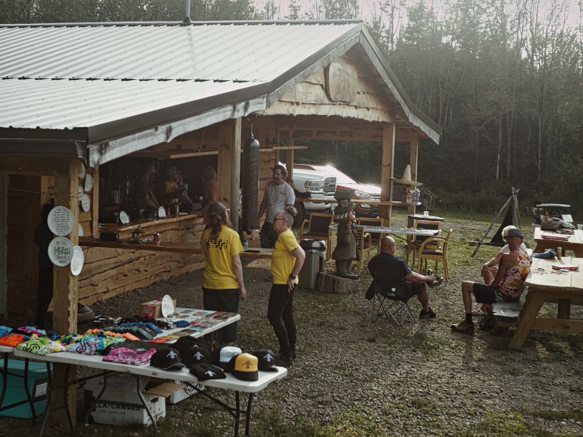Rustic open-air wooden bar with a corrugated metal roof, set against a backdrop of dense trees with hazy light breaking through. In the foreground, a folding table holds black snapback hats, colorful merchandise, and a cardboard box; two people in matching yellow T-shirts stand chatting near the bar, while two others sit in camp chairs and at a picnic table to the right. A red pickup truck is parked just behind the structure, and a chainsaw-carved bear sculpture stands near the center.