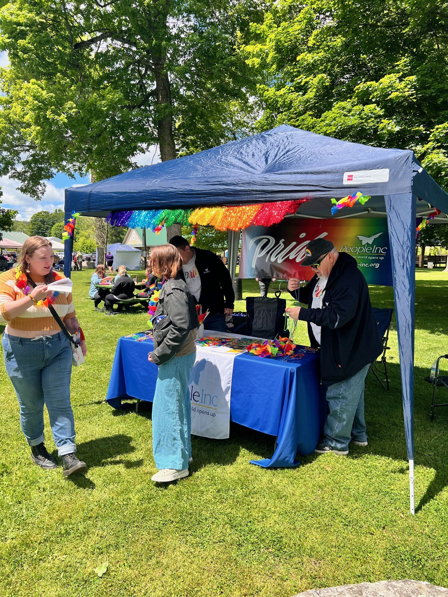 A navy pop-up canopy tent decorated with rainbow fringe and tinsel at an outdoor Pride event on a sunny green lawn, with a blue-draped table below bearing a "People Inc — the world opens up" banner and colorful paper crafts spread across it. A woman in a striped orange sweater stands to the left reviewing materials, another attendee in a gray jacket browses the table, and an older man in black assists from the other side, with tall leafy trees filling the bright background.