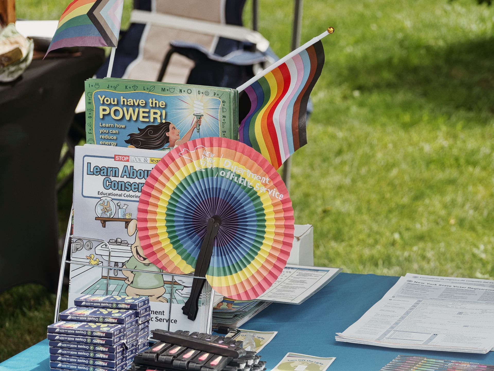 Outdoor tabling setup on a blue tablecloth, centered around a rainbow paper fan printed with "New York Department of Public Service." Behind it, pamphlets including "You have the POWER! Learn how you can reduce energy" and a partially visible "Learn About Conservation" educational coloring book are displayed in a rack, flanked by two Progress Pride flags. Stacks of Prang crayon boxes and pens sit in the foreground left, while New York State voter registration forms are fanned out on the right, with green grass and a folding chair blurred in the background.