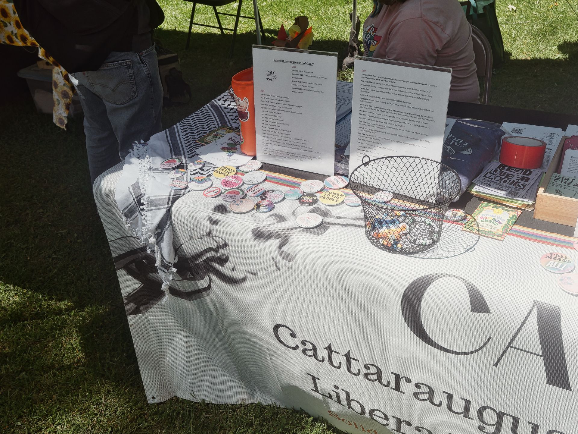 An outdoor tabling setup for the Cattaraugus Libera[tion] — the full name cut off by the frame — with a white tablecloth printed with broken chains and the organization's "CAL" logo. The table holds scattered activist pinback buttons, a wire basket filled with more buttons, two acrylic-framed informational documents titled "Important Events Timeline of CALC," an orange bucket, and a stack of books including titles reading "DEAD QUEER BOOKS" and "DIRT CHEAP" to the right. A keffiyeh is draped across the left side of the table, and partial figures of attendees are visible in the background on grass.