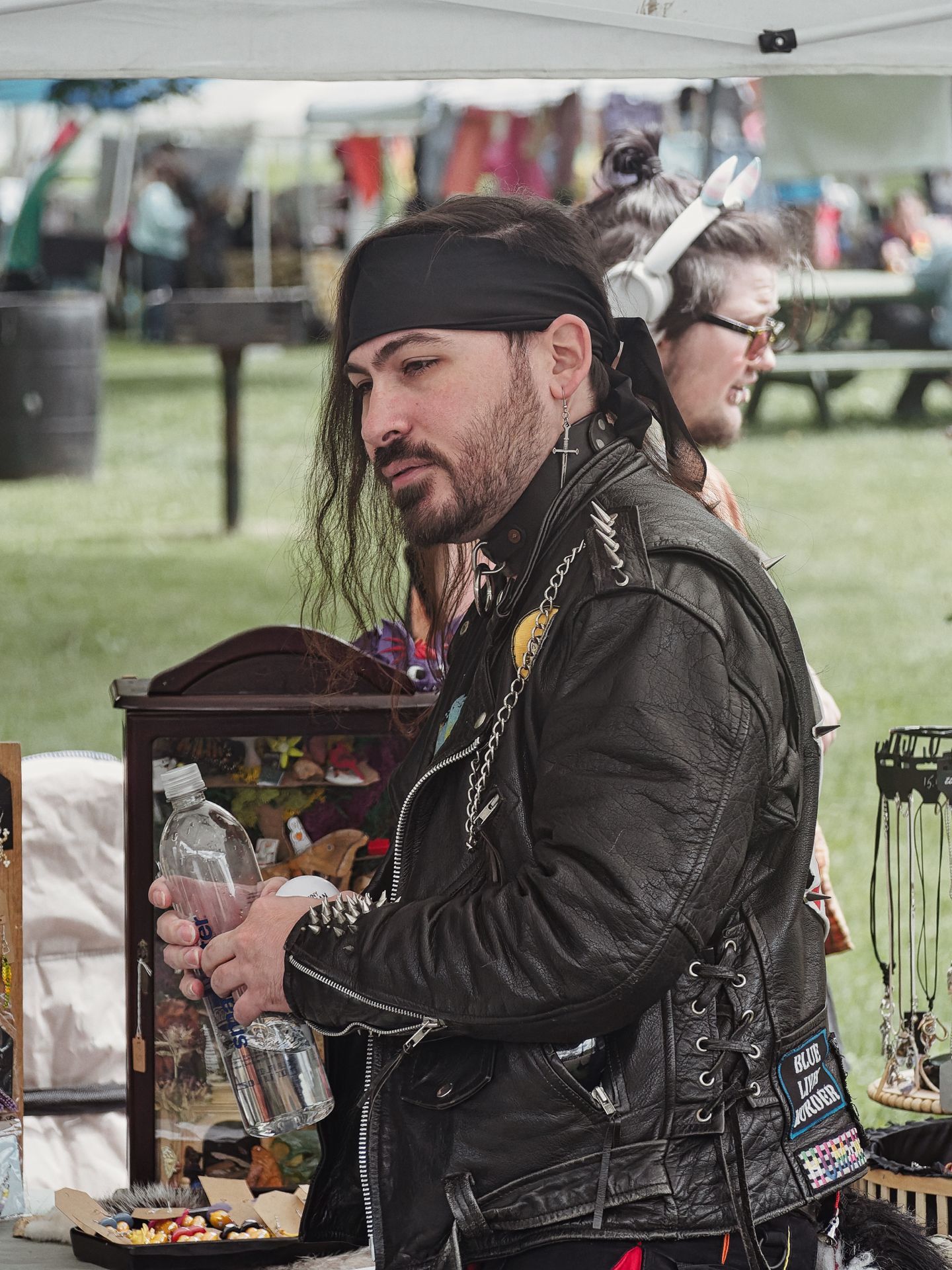 Man in a spiked black leather vest covered in pins and chains, wearing a black bandana and cross earring, holding a clear plastic water bottle at what appears to be an outdoor vendor booth. Behind him, a second person wearing cat-ear headphones is partially visible, with a small wooden display case, jewelry rack, and festival tents blurred in the background.