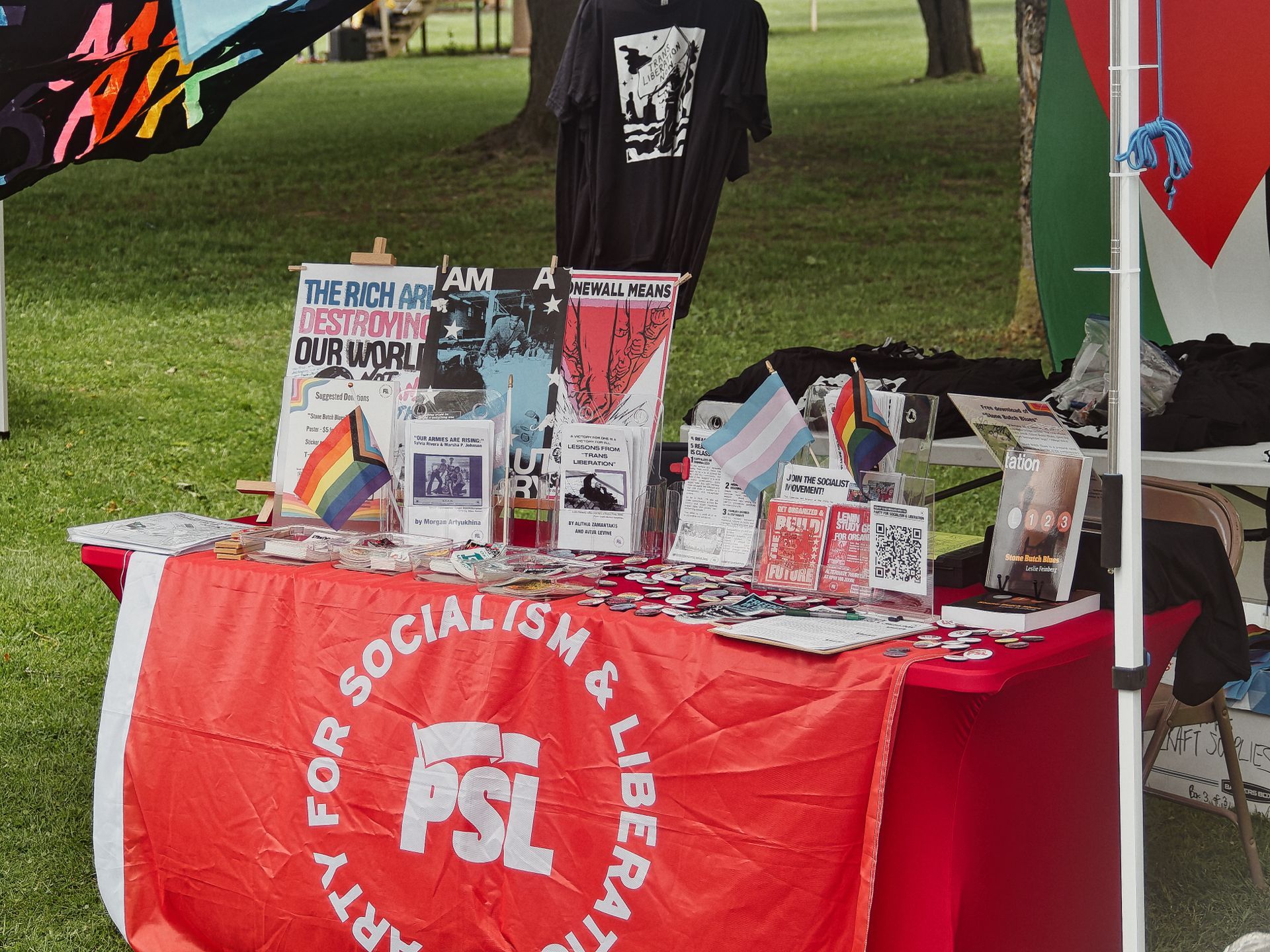 An outdoor tabling setup draped in a red banner reading "PSL — PARTY FOR SOCIALISM & LIBERATION," covered with pins, pamphlets, and small pride and transgender flags. Posters in the background read "THE RICH ARE DESTROYING OUR WORLD," "STONEWALL MEANS," and "LESSONS FROM TRANS LIBERATION," with a black graphic T-shirt hanging above; a grassy park and trees are visible behind the booth.