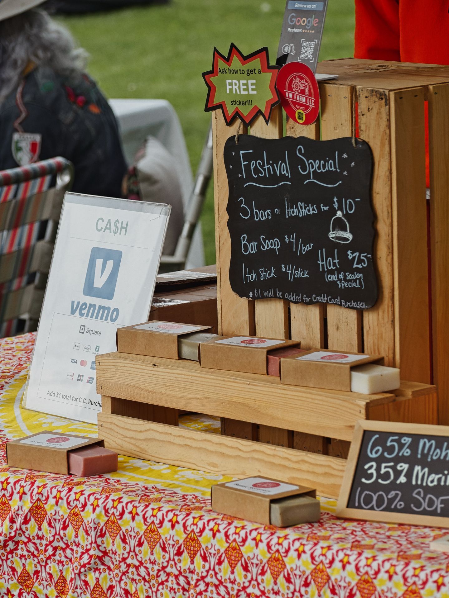 Outdoor market vendor table covered in a red-and-yellow floral cloth, displaying bars of soap arranged on a tiered wooden crate display. A chalkboard sign reads "Festival Special! 3 bars or Itch Sticks for $10 — Bar Soap $4/bar, Itch Stick $4/stick, Hat $25 (end of season special) — $1 will be added for Credit Card Purchases," with a red starburst tag above reading "Ask how to get a FREE sticker!!!" and a Google Reviews sign above that; to the left, a printed sign reads "CA$H / venmo / Square / Add $1 total for C.C. Purcha—"; a smaller chalkboard in the foreground right reads "65% Moh— / 35% Meri— / 100% SOF—," partially cut off.
