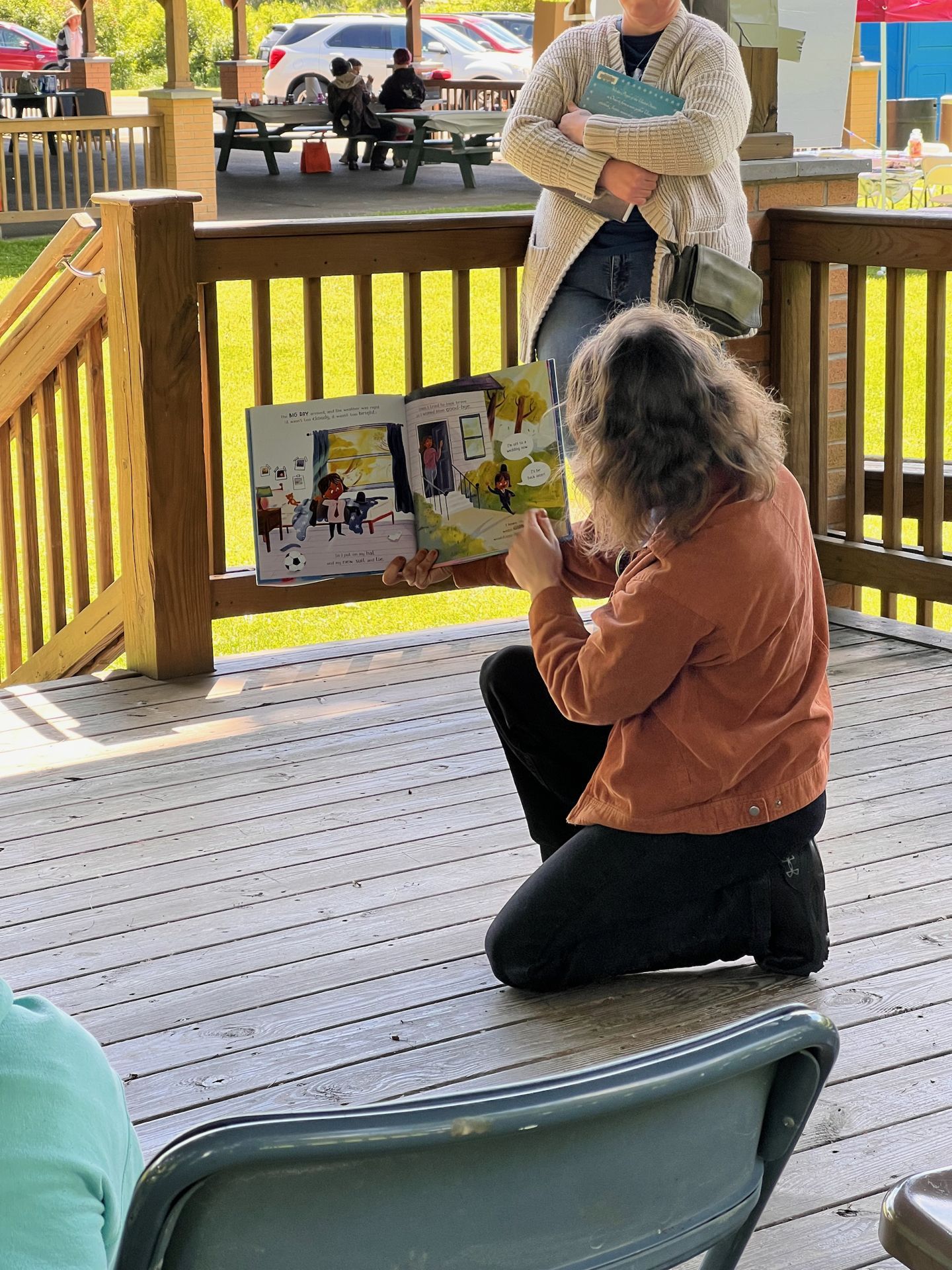 Person in an orange jacket kneeling on a wooden deck, holding open a colorful illustrated children's book toward an unseen audience. Behind them, another adult in a cream cardigan stands with arms crossed holding a teal booklet; picnic tables and parked cars are visible in the sunny background.