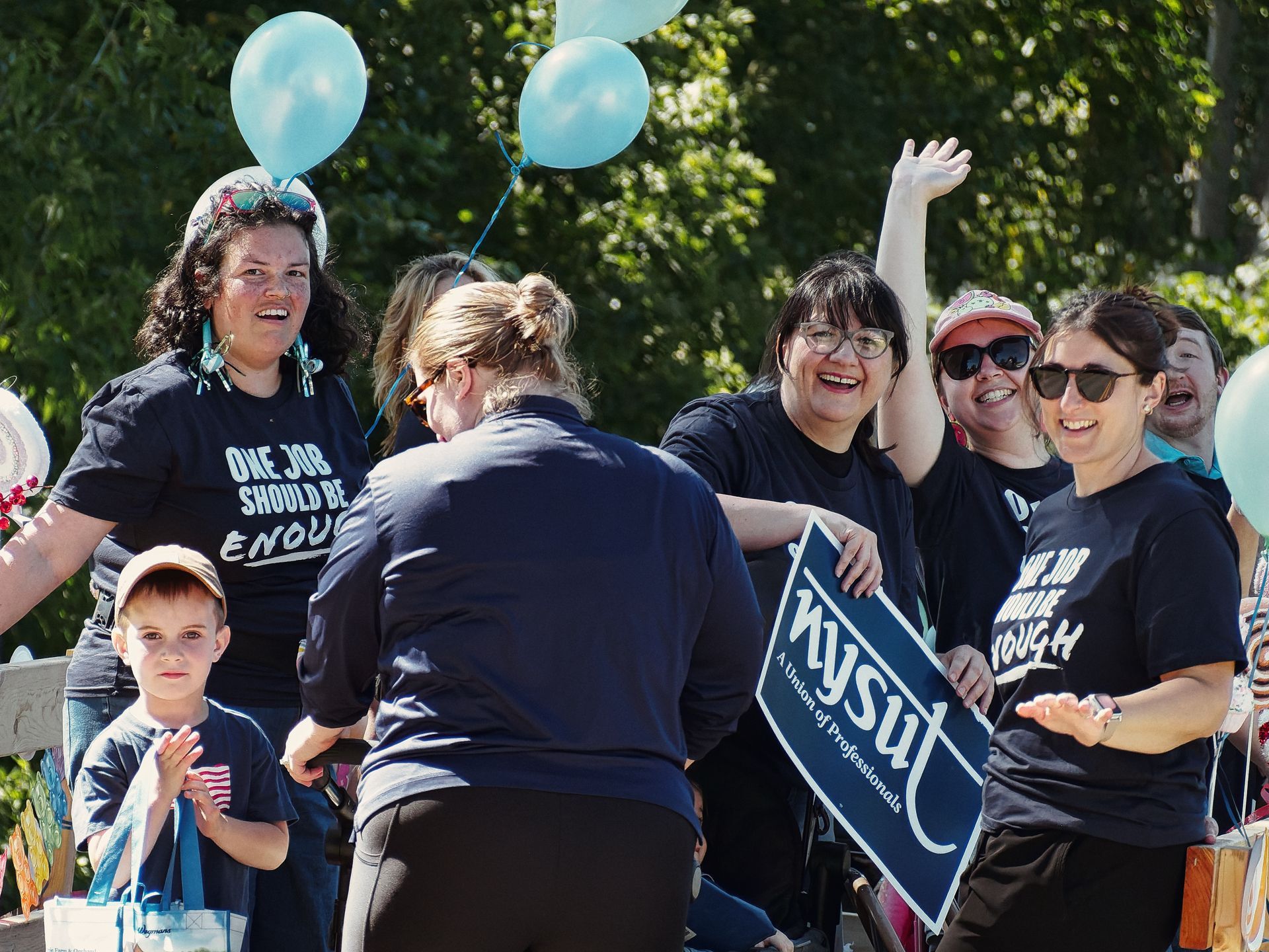A cheerful group of adults and one young child at what appears to be a Labor Day parade, several wearing black T-shirts reading "ONE JOB SHOULD BE ENOUGH." One woman holds a navy blue sign reading "NYSUT — A Union of Professionals," another waves with her hand raised, and light blue balloons float above the crowd against a backdrop of sunlit green trees.