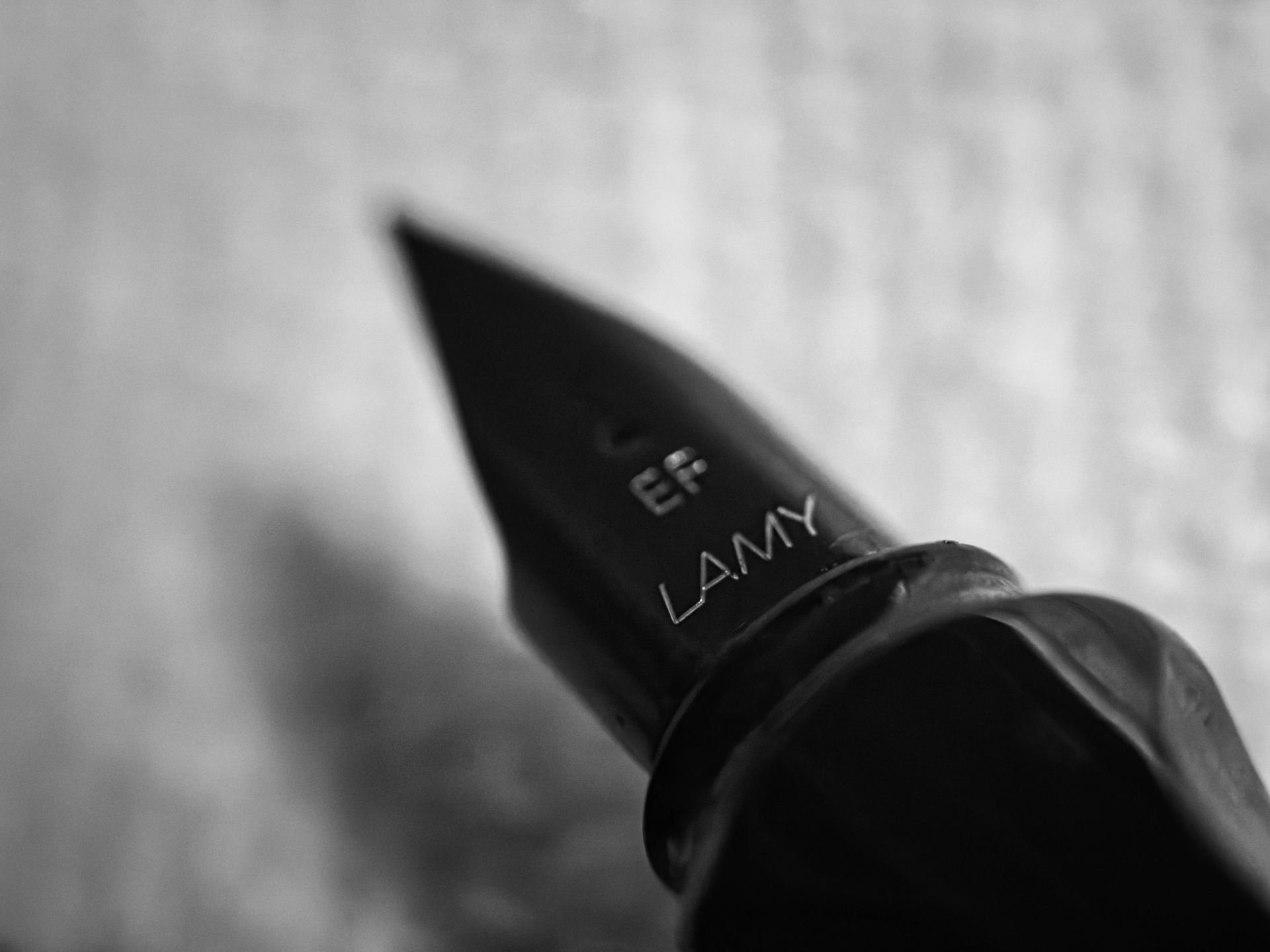 Black-and-white extreme close-up of a matte black fountain pen nib engraved with "EF" and "LAMY," angled diagonally toward the upper left, its tip sharp against a softly blurred, textured light background.