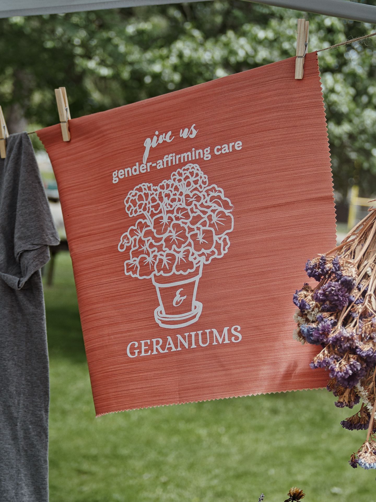 A terracotta-orange fabric panel clipped to a clothesline with wooden pegs, printed in white with the text "give us gender-affirming care & GERANIUMS" surrounding an illustrated potted geranium plant. A gray t-shirt hangs to the left, dried lavender bundles hang to the right, and lush green trees blur softly in the background.