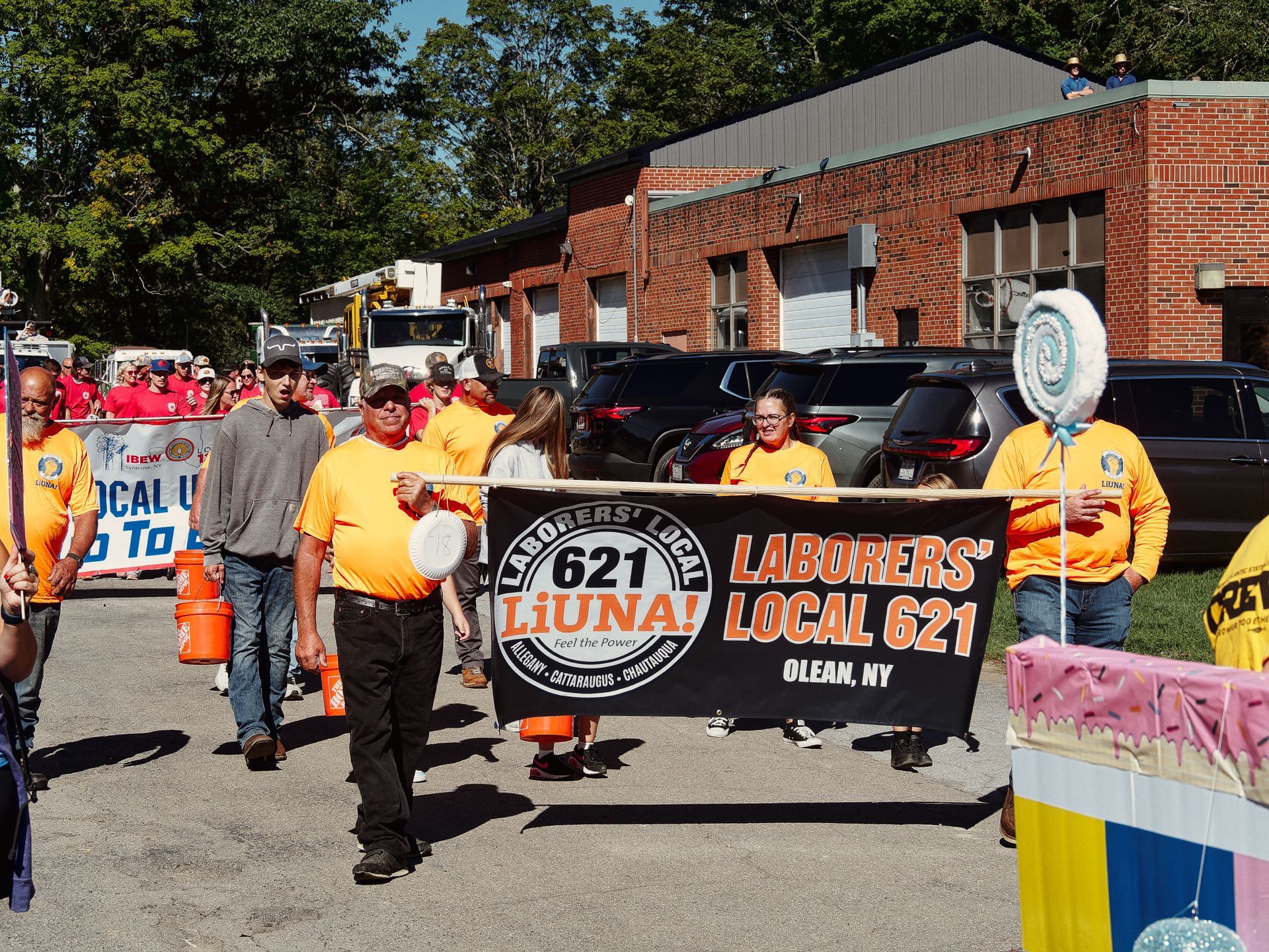 Workers in bright orange LiUNA shirts march down a street carrying a black banner reading "LABORERS' LOCAL 621 LiUNA! Feel the Power — Allegany · Cattaraugus · Chautauqua / LABORERS' LOCAL 621 OLEAN, NY." Behind them, a crowd in red shirts follows alongside an IBEW banner, with a brick fire station and parked vehicles in the background.