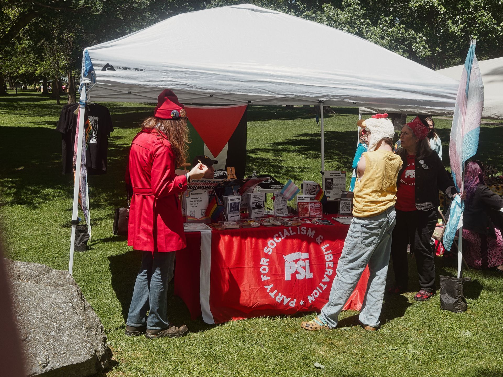 Outdoor tabling event in a sunny park, centered on a white canopy tent sheltering a table draped in a red banner reading "PARTY FOR SOCIALISM & LIBERATION — PSL." A person in a long red coat and red beret stands at the left side of the table, facing two others — one in a yellow vest and fuzzy white hat, one in a black shirt with a red bandana — on the right. A Palestinian flag hangs at the back of the table, a transgender pride flag flies on the far right, and a rack of black t-shirts hangs at the left. The table surface is covered with pamphlets, buttons, and small printed materials.