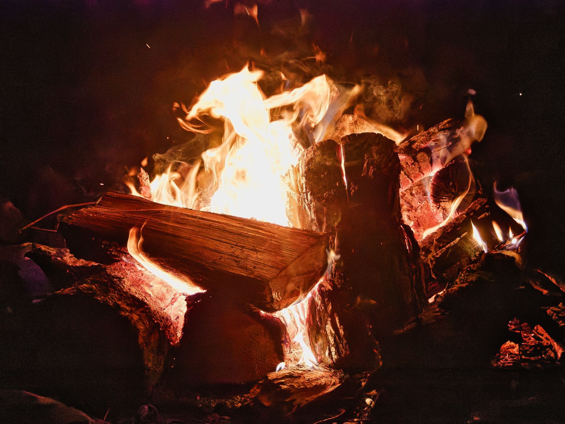 A campfire burning at night, with split logs and thick rounds of wood stacked loosely around a blazing white-yellow core, flames curling upward against a pitch-black background, embers glowing deep red along the charred wood edges.