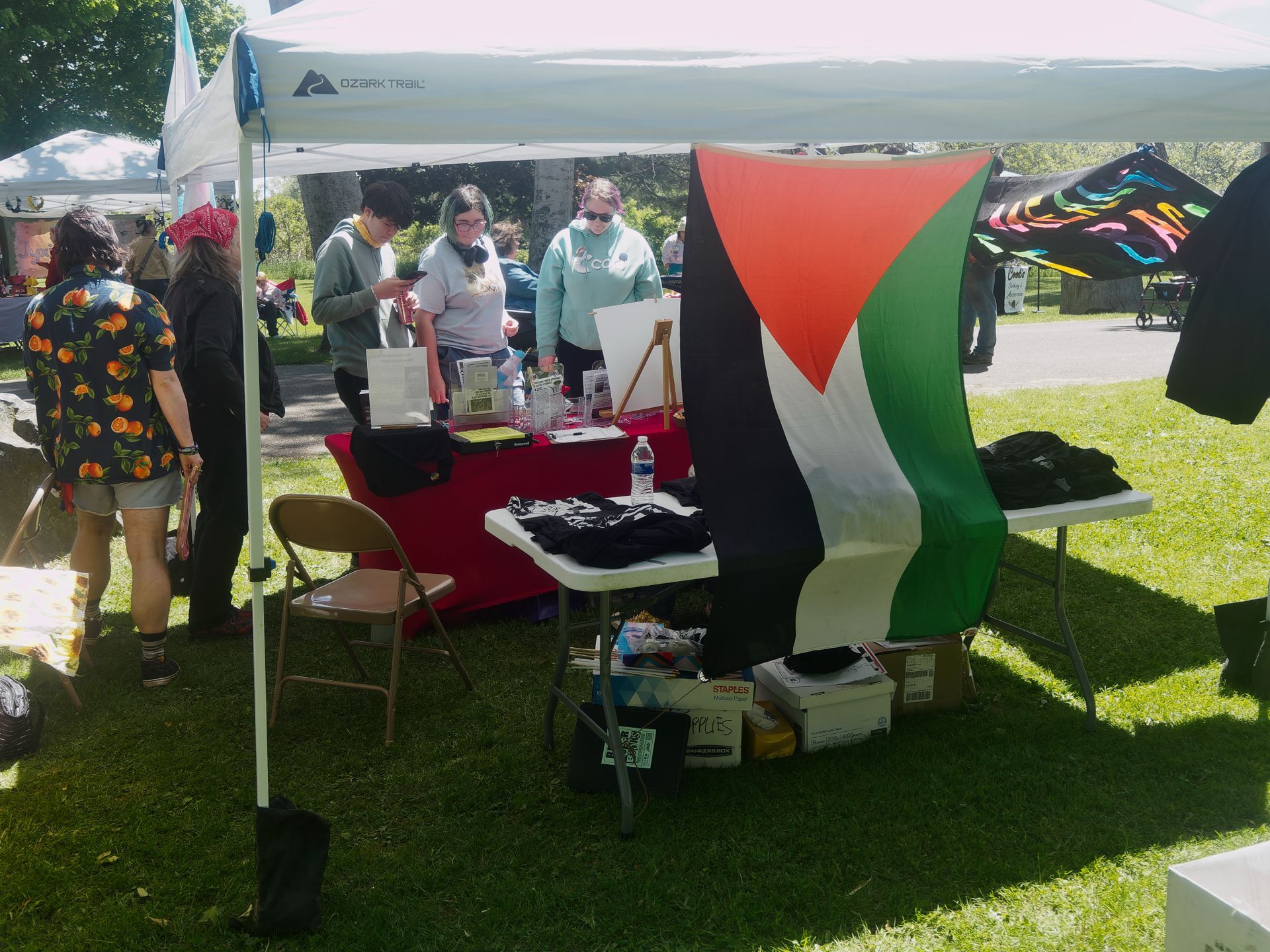 An outdoor vendor booth under an Ozark Trail canopy on a sunny grass lawn, with a large Palestinian flag — black, white, red, and green — draped prominently at the center. Three people browse items on red-and-white-tableclothed tables stacked with folded black shirts, pamphlets, and a water bottle, while a fourth person in an orange-print shirt walks past on the left, and cardboard boxes labeled "SUPPLIES" sit on the ground beneath the tables.