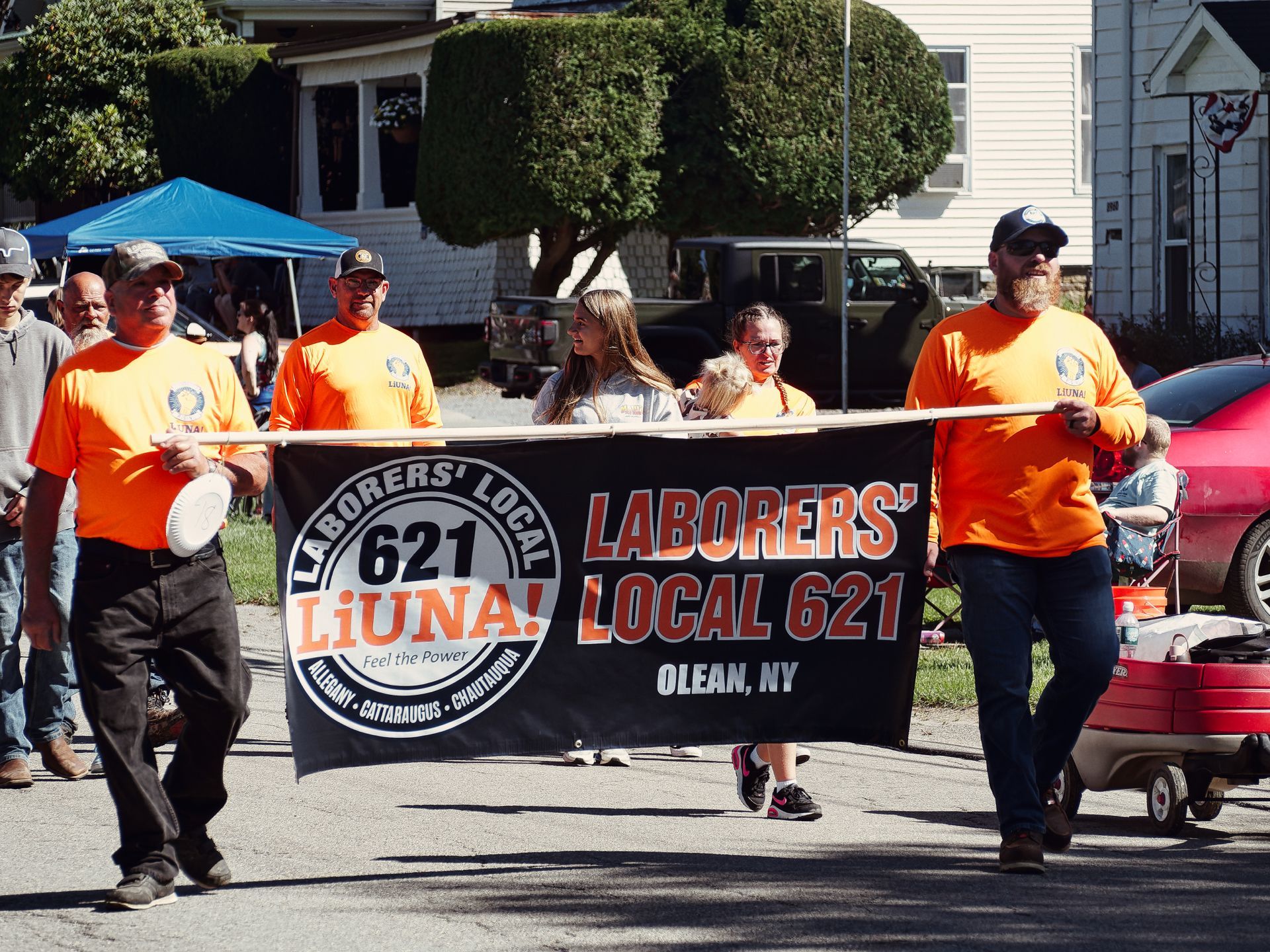 Group of people in matching orange LiUNA shirts marching down a residential street, carrying a large black banner reading "Laborers' Local 621 · LiUNA! · Feel the Power · Allegany · Cattaraugus · Chautauqua" on the left and "Laborers' Local 621 · Olean, NY" on the right, with a blue pop-up tent, parked trucks, and white clapboard houses visible in the background.