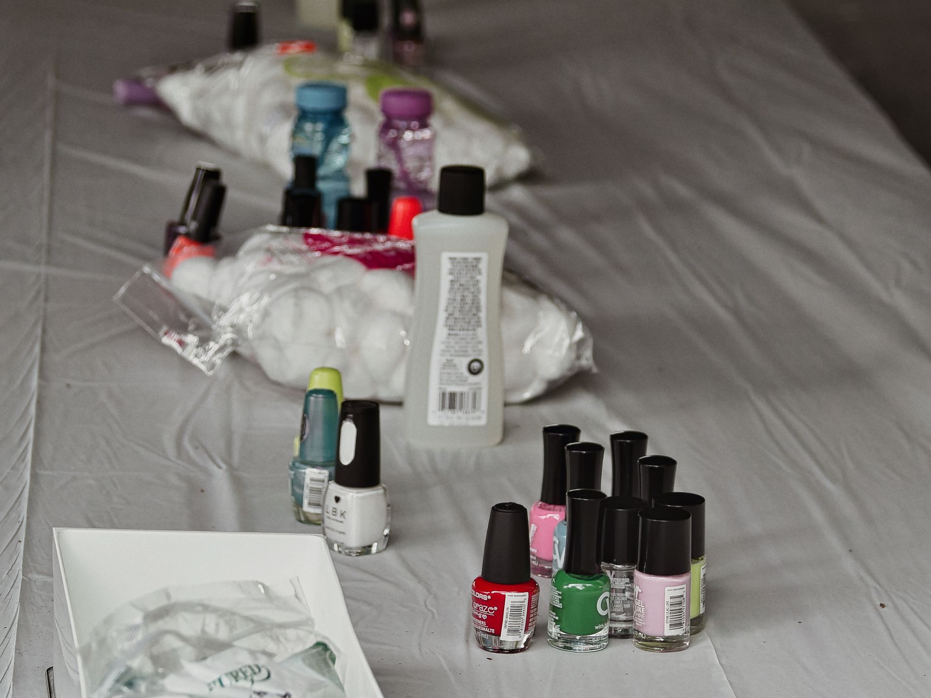Nail polish bottles in red, green, pink, white, and teal clustered in the foreground on a white plastic-covered table, alongside a clear lotion bottle turned label-out, bags of cotton balls, and more polish bottles receding into the blurred background.