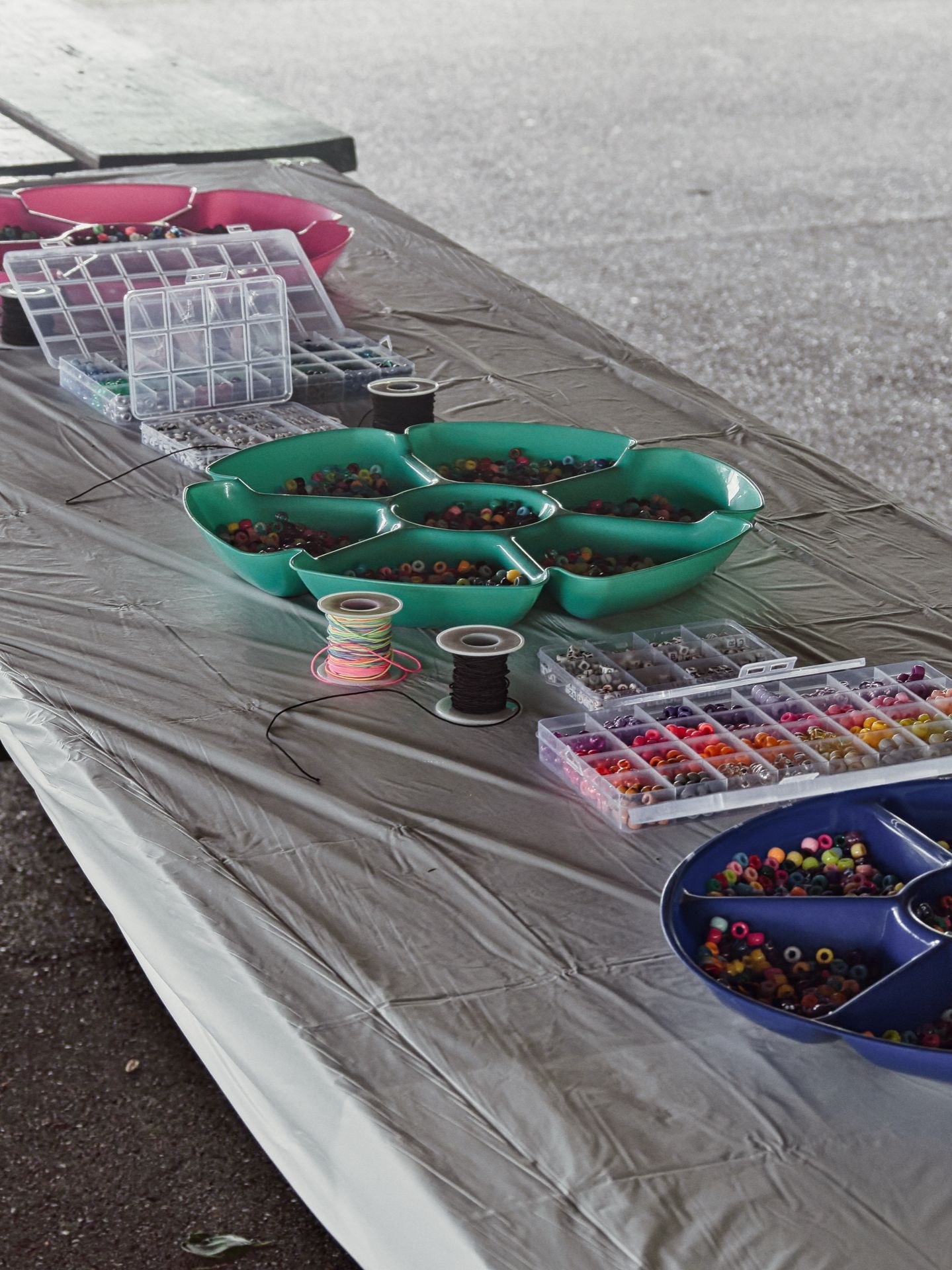 A folding table covered in a gray plastic tablecloth holds a craft bead station: a teal multi-section tray and a navy divided bowl overflow with colorful pony beads, while clear plastic grid organizers sorted by bead color and size flank them, and two spools of elastic cord — one multicolored, one black — sit in the center foreground.