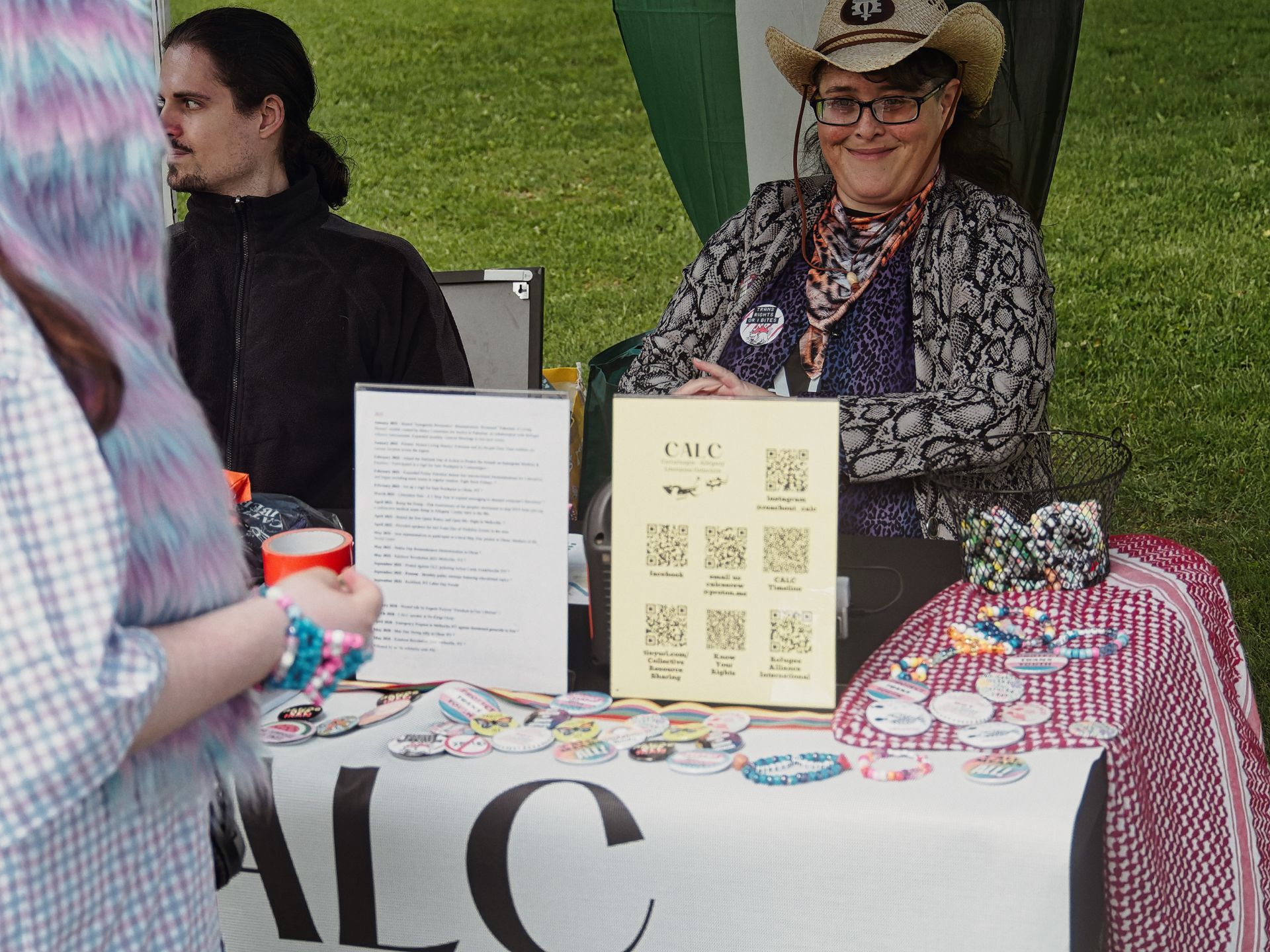 Person in a straw cowboy hat, glasses, and a snakeskin-print jacket smiling warmly behind an outdoor event table labeled "ALC" in large letters on a white tablecloth, covered with colorful buttons, beaded bracelets, and a yellow QR code sign reading "CMC" with links to Instagram, Facebook, email, and resources including "Know Your Rights" and "Refugee Alliance International." A second person in a black zip-up jacket sits to the left, gazing away, while a visitor with pink-and-blue hair stands blurred in the foreground right. Green grass is visible in the background.
