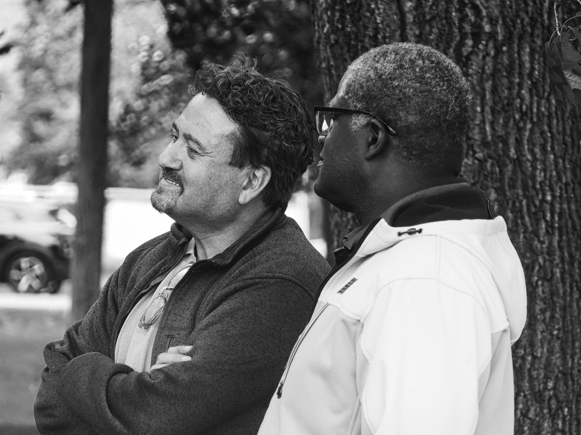 Black-and-white shot of two men standing near a large tree trunk on the right — the man on the left, curly-haired with a goatee and arms crossed, smiles as he looks toward the other man, who wears glasses and a white zip-up hoodie and gazes upward. A parked car is softly blurred in the shallow depth of the background at left.