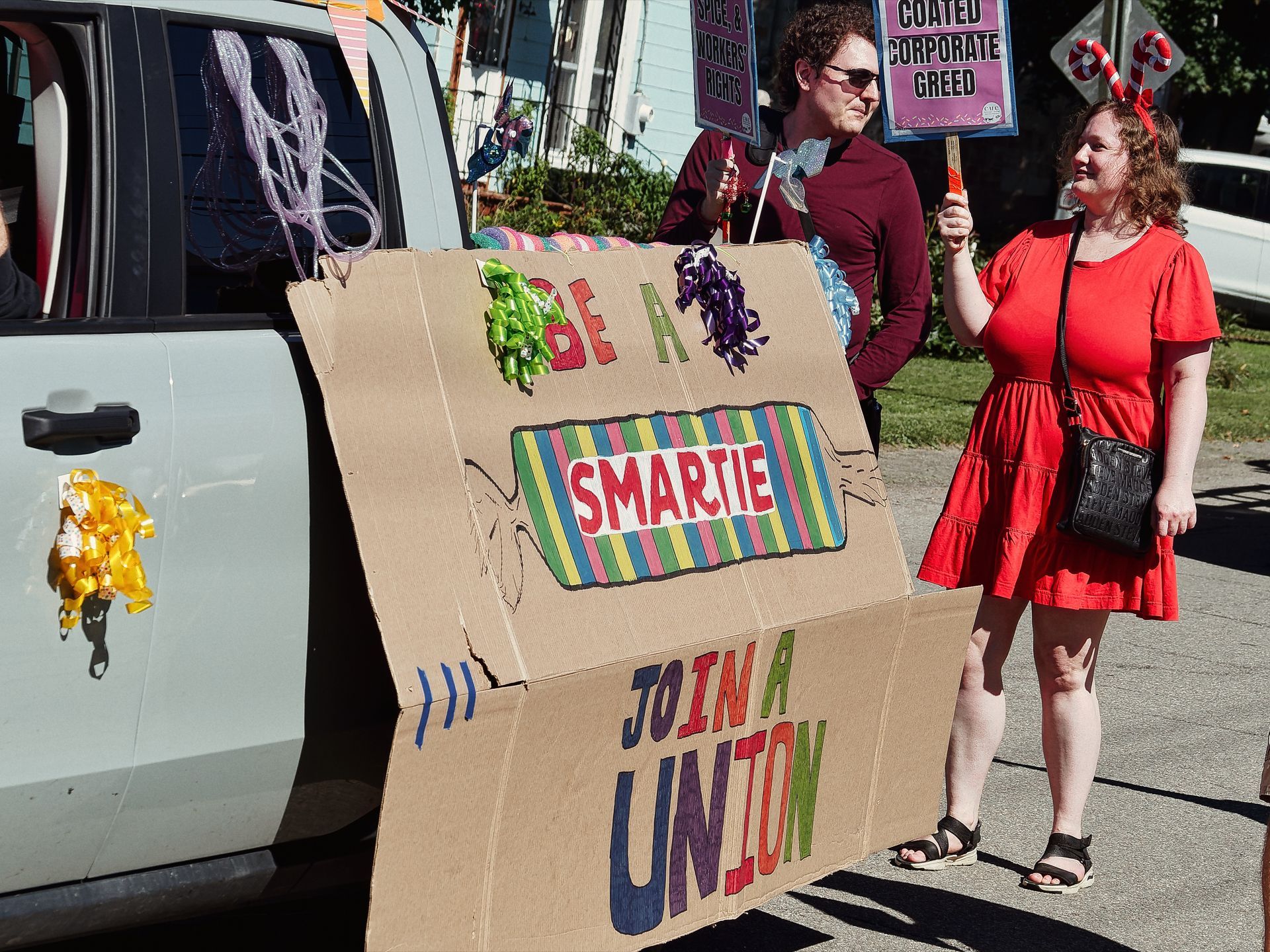 Two protesters stand on a street beside a large handmade cardboard sign reading "BE A SMARTIE — JOIN A UNION," decorated with colorful ribbon bows and a hand-drawn Smarties candy wrapper. The woman on the right wears a red dress and candy cane headband, holding a sign partially visible reading "COATED CORPORATE GREED," while the person to her left in a dark red top holds additional protest materials; a white SUV with a gold ribbon decoration is parked to the left.