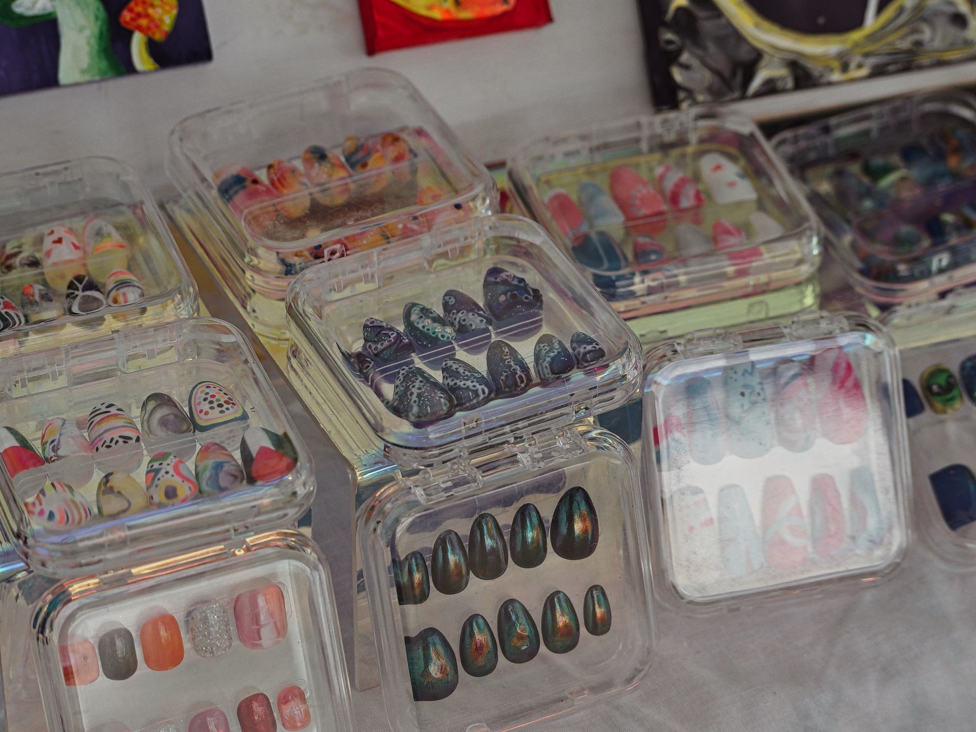 Rows of clear plastic clamshell cases displaying sets of press-on nails in various painted designs — deep iridescent green-black almond shapes, dark purple with leopard print, pastel watercolor swirls, and peach-pink gradients — arranged across a white surface at what appears to be a vendor table, with colorful artwork hanging on the wall blurred in the background.