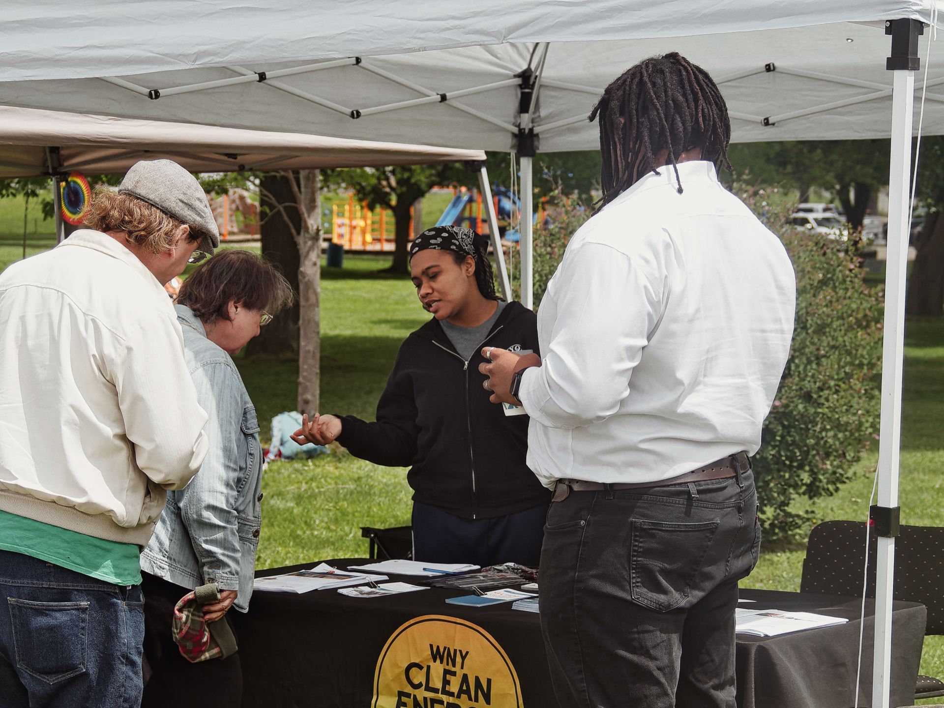 Four people gathered under a white pop-up canopy at an outdoor event booth draped in a black tablecloth bearing a yellow circular logo reading "WNY CLEAN ENER—" (partially obscured). A person in a black hoodie and bandana headband stands behind the table, gesturing while speaking to two attendees on the left — one in a gray flat cap and cream jacket, one in a denim jacket — while a fourth person in a white button-down and locs stands to the right. A park playground with a slide is visible blurred in the background.