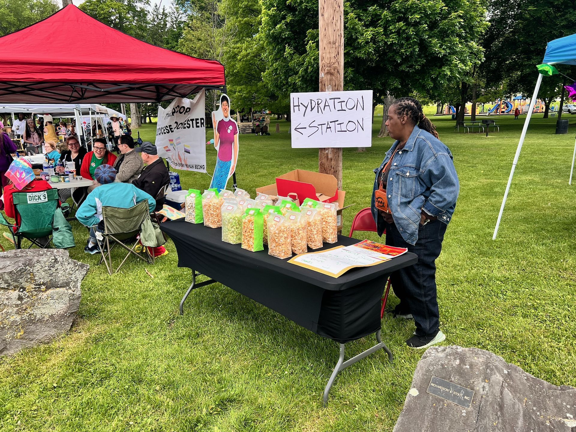 A woman in a denim jacket with locs stands beside a black-draped folding table lined with bagged popcorn at an outdoor community event, next to a handwritten sign reading "Hydration ← Station." Behind her, a life-size cardboard cutout and a banner partially reading "STOP ROCHESTER" stand near a red canopy tent where several people sit in camp chairs on a grassy park lawn.
