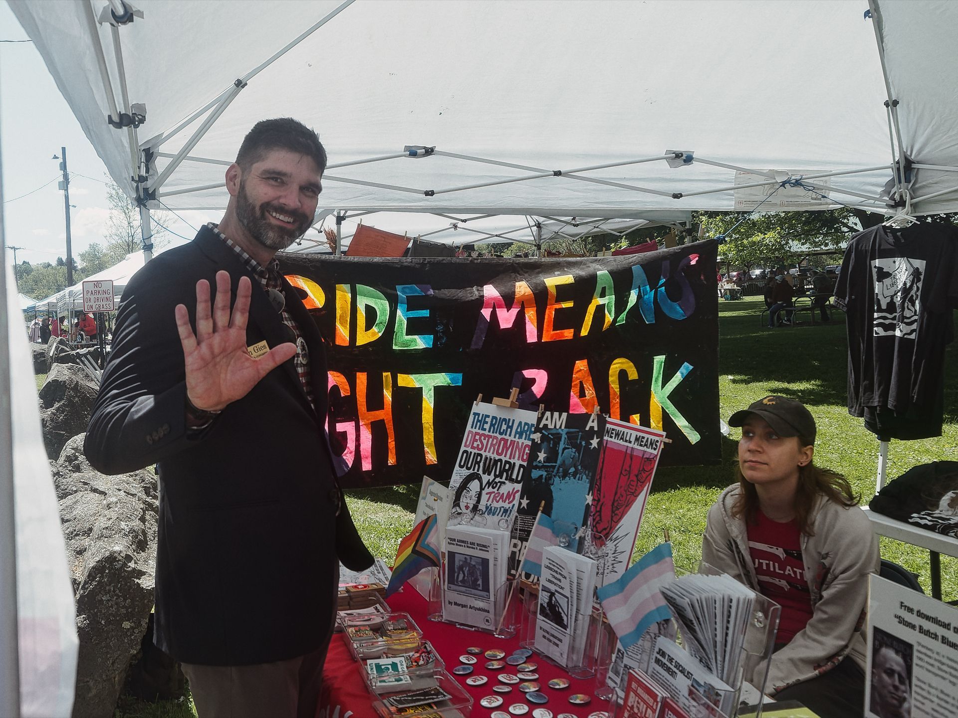 An outdoor Pride event booth under a white canopy, with a smiling man in a black blazer raising his hand toward the camera on the left, and a woman in a baseball cap seated behind a red table stacked with zines, buttons, pins, and a small transgender pride flag. A large black banner behind them reads "PRIDE MEANS FIGHT BACK" in rainbow letters, and a pamphlet on the table reads "THE RICH ARE DESTROYING OUR WORLD NOT TRANS YOUTH!" Black T-shirts hang from the canopy frame on the right, and green parkland stretches into the background.