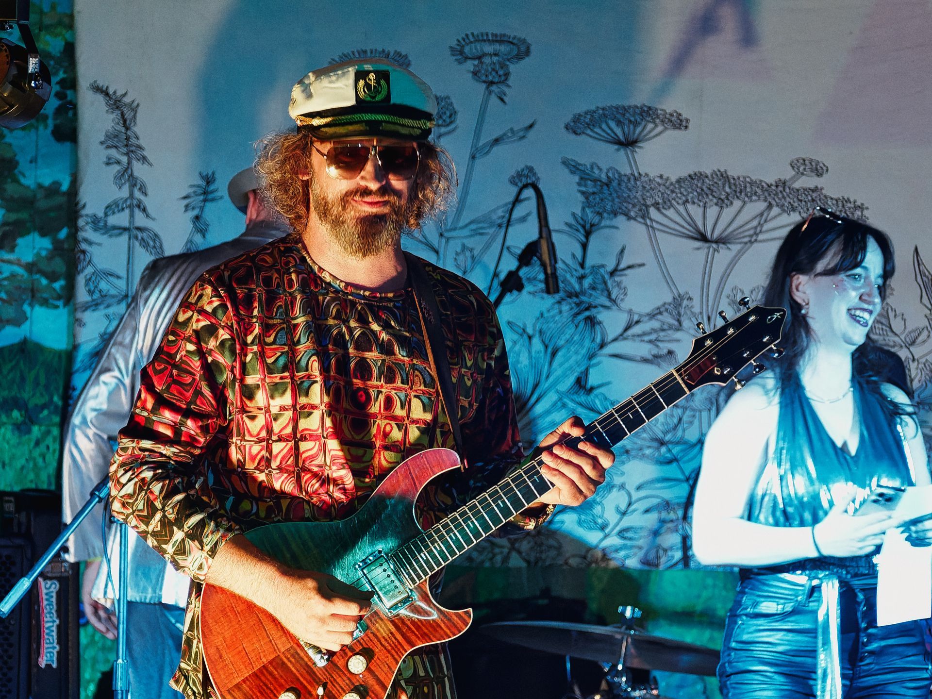 A bearded guitarist in a psychedelic patterned shirt, captain's hat, and tinted aviator sunglasses plays a flame-maple electric guitar at center stage, grinning broadly. To his right, a woman in a metallic blue outfit laughs, while a third performer in white stands partially visible at the far left, all backed by a blue-lit botanical line-art mural. A mic stand and a "Sweetwater" branded amp are visible in the lower left foreground.