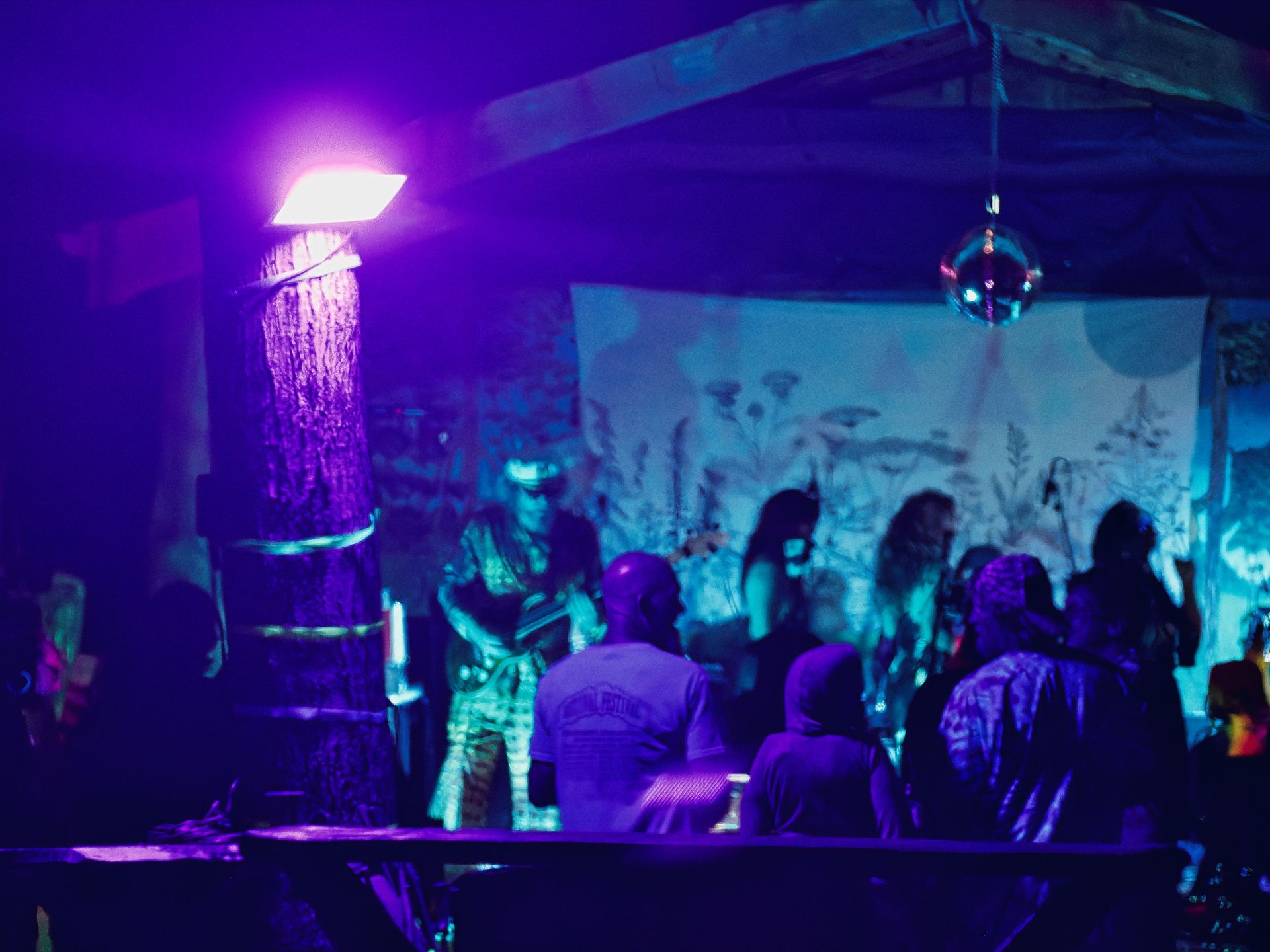 Crowd gathered in front of a makeshift outdoor stage bathed in deep purple light, with a performer in a reflective outfit near a tree trunk mounted with a bright pink-white floodlight on the left. A hand-painted botanical backdrop hangs behind the stage, and a small disco ball dangles from a wooden beam overhead.