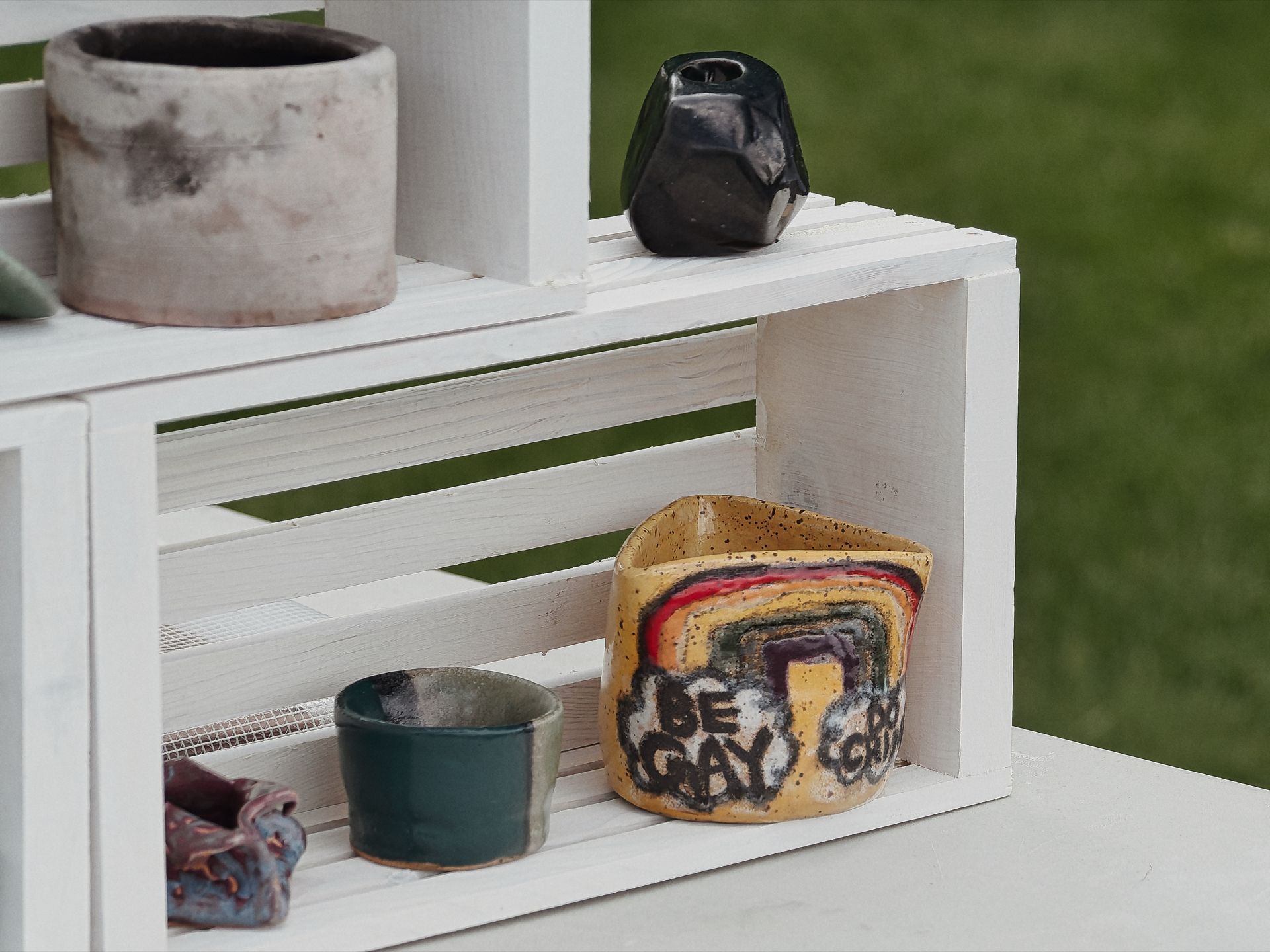 Handmade ceramic pieces displayed on white painted wood crate shelving, set against a blurred green grass background. On the lower shelf sits a teal-glazed bowl and a yellow, irregularly shaped vessel painted with rainbow-bordered text reading "BE GAY DO GAY"; above on the upper shelf, a matte grey cylindrical pot sits upper left and a small glossy black angular vase sits upper right.