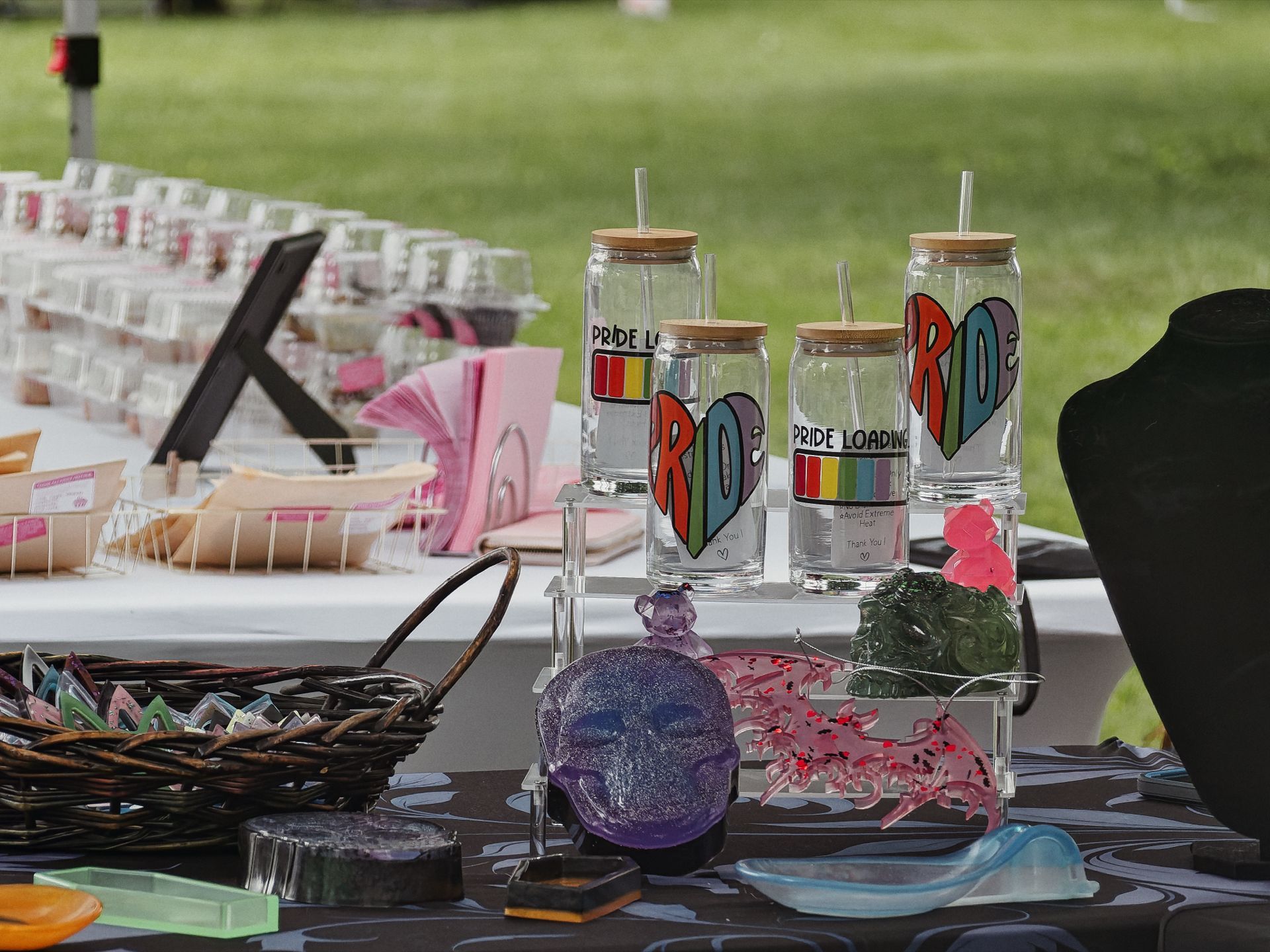 Outdoor vendor table covered in handmade goods, centered on four glass can-style cups with bamboo lids and straws printed with "PRIDE LOADING" and a rainbow heart design. In the foreground, a wicker basket holds small trinkets alongside a glittery purple skull figurine, a pink acrylic lobster, a dark green resin piece, and scattered colorful objects; rows of packaged items stretch blurred into the background against a green lawn.