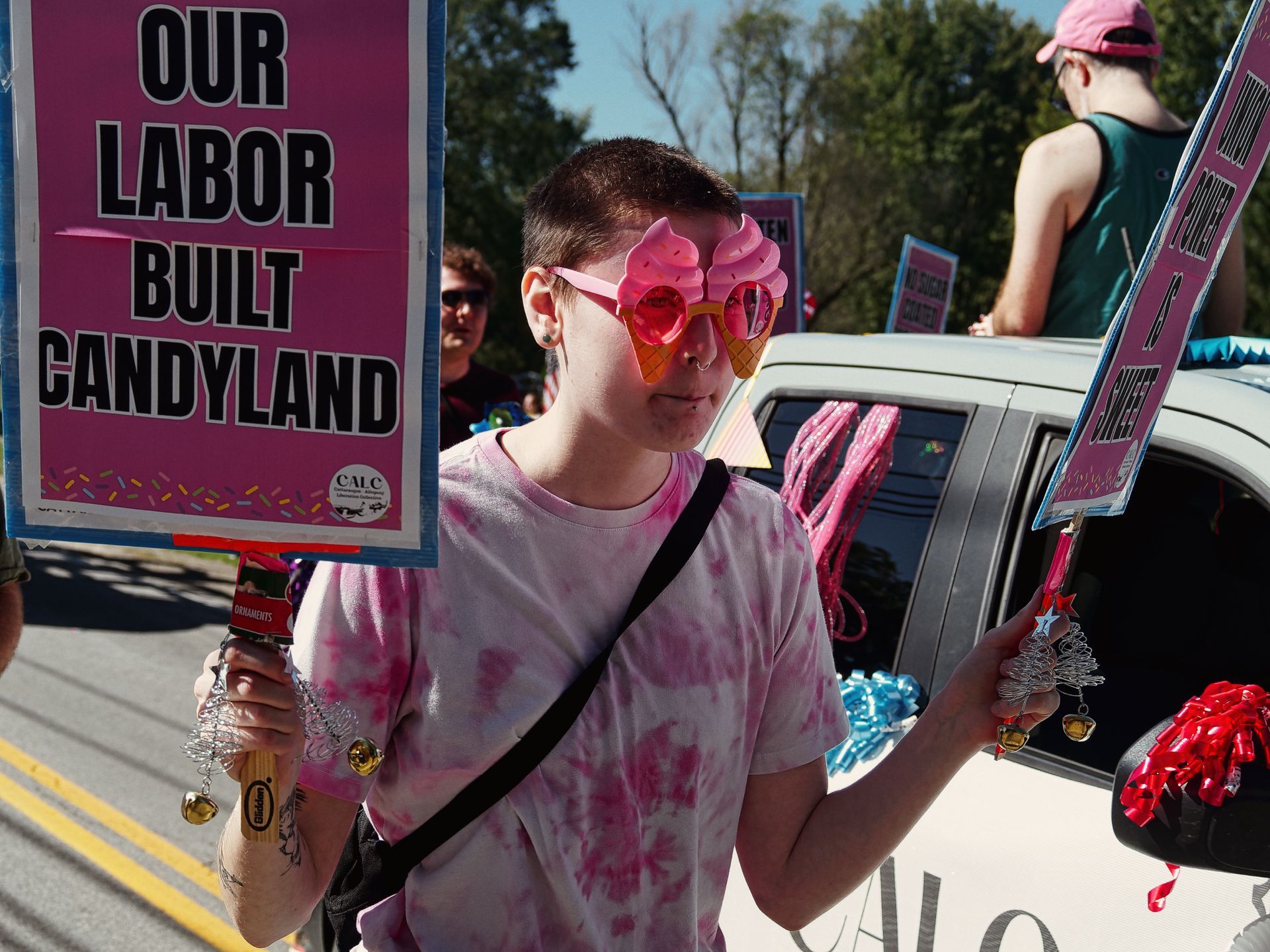 Person in a pink tie-dye shirt and oversized pink ice cream cone novelty sunglasses holding a hot-pink protest sign reading "OUR LABOR BUILT CANDYLAND" with a CALC logo at the bottom, marching alongside a decorated white vehicle during what appears to be a labor parade. A second pink sign partially visible on the right reads "UNION POWER IS SWEET," and a person in a pink cap sits atop the vehicle in the background.