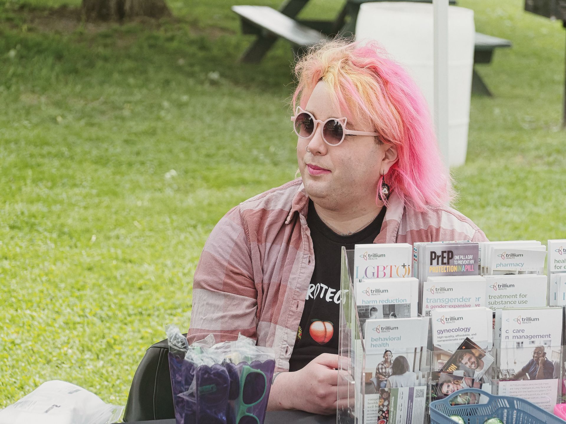 Person with pink-and-yellow hair and round pink sunglasses sitting behind an outdoor health resource table, wearing a black "PROTECT" T-shirt with a peach emoji and an open pink flannel. The table holds a clear acrylic brochure stand filled with Trillium Health pamphlets labeled "PrEP," "harm reduction," "transgender & gender-expansive," "behavioral health," "gynecology," "treatment for substance use," and "care management," with a basket of supplies to the right; a grassy park and picnic tables are blurred in the background.