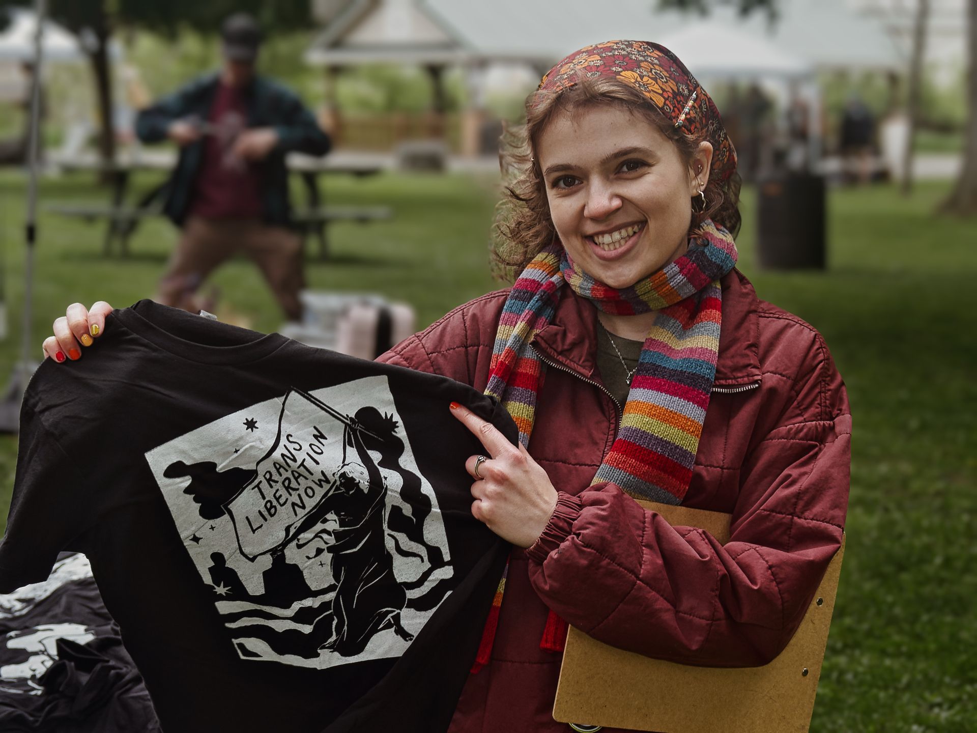 Smiling person in a dark red quilted jacket, rainbow striped scarf, and floral headband holds up a black t-shirt printed with a silhouetted figure raising a banner that reads "TRANS LIBERATION NOW," pointing to it with a ringed finger. A green park lawn, picnic tables, and a gazebo are blurred in the background, with another person standing at a table to the left.