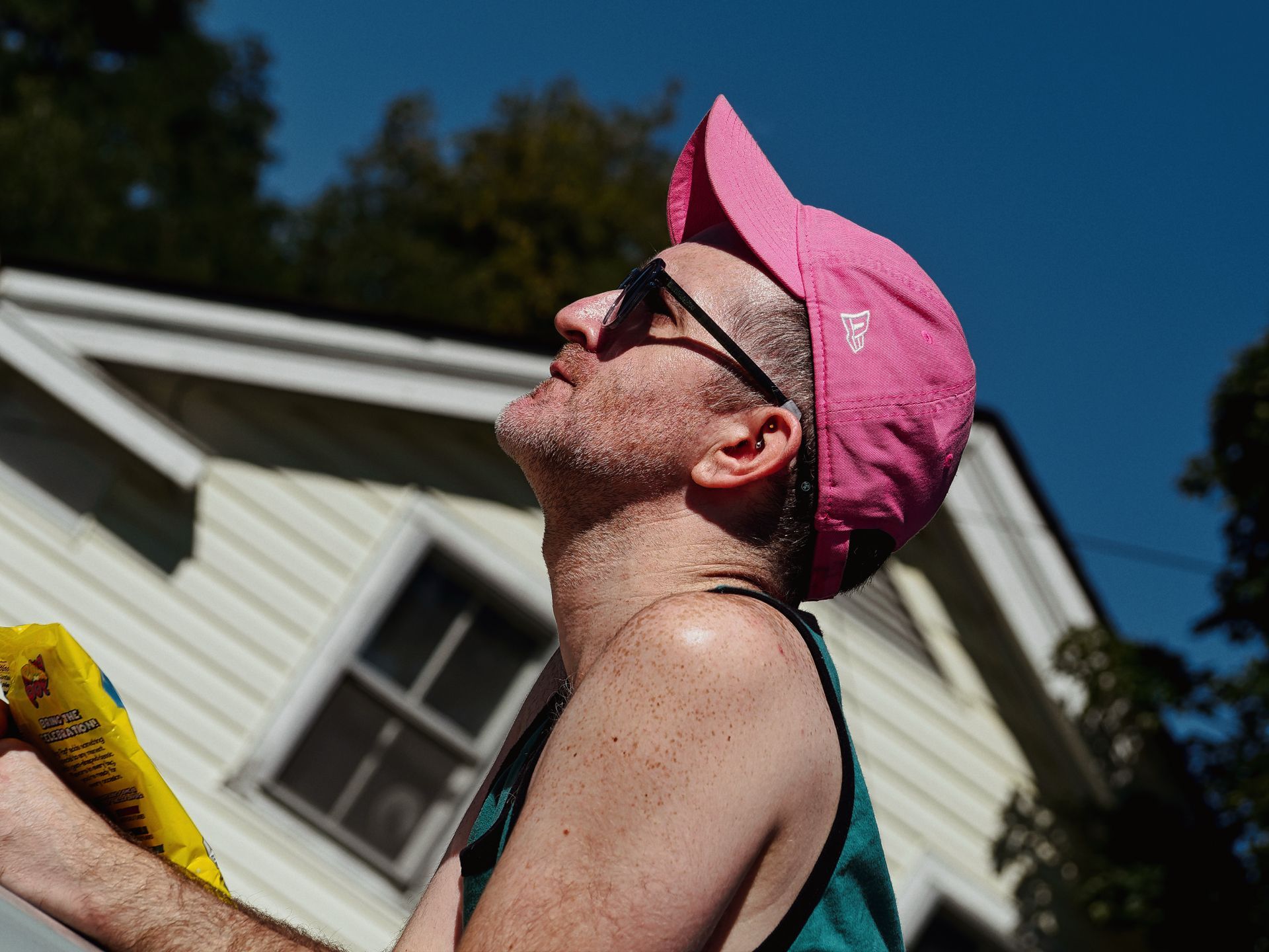 Man in a bright pink running cap and black sunglasses tilting his head back toward a deep blue sky, shot from a low angle that makes him dominate the frame. He's wearing a teal tank top and holding a yellow snack bag in his left hand; a white clapboard house and green trees blur softly in the background.