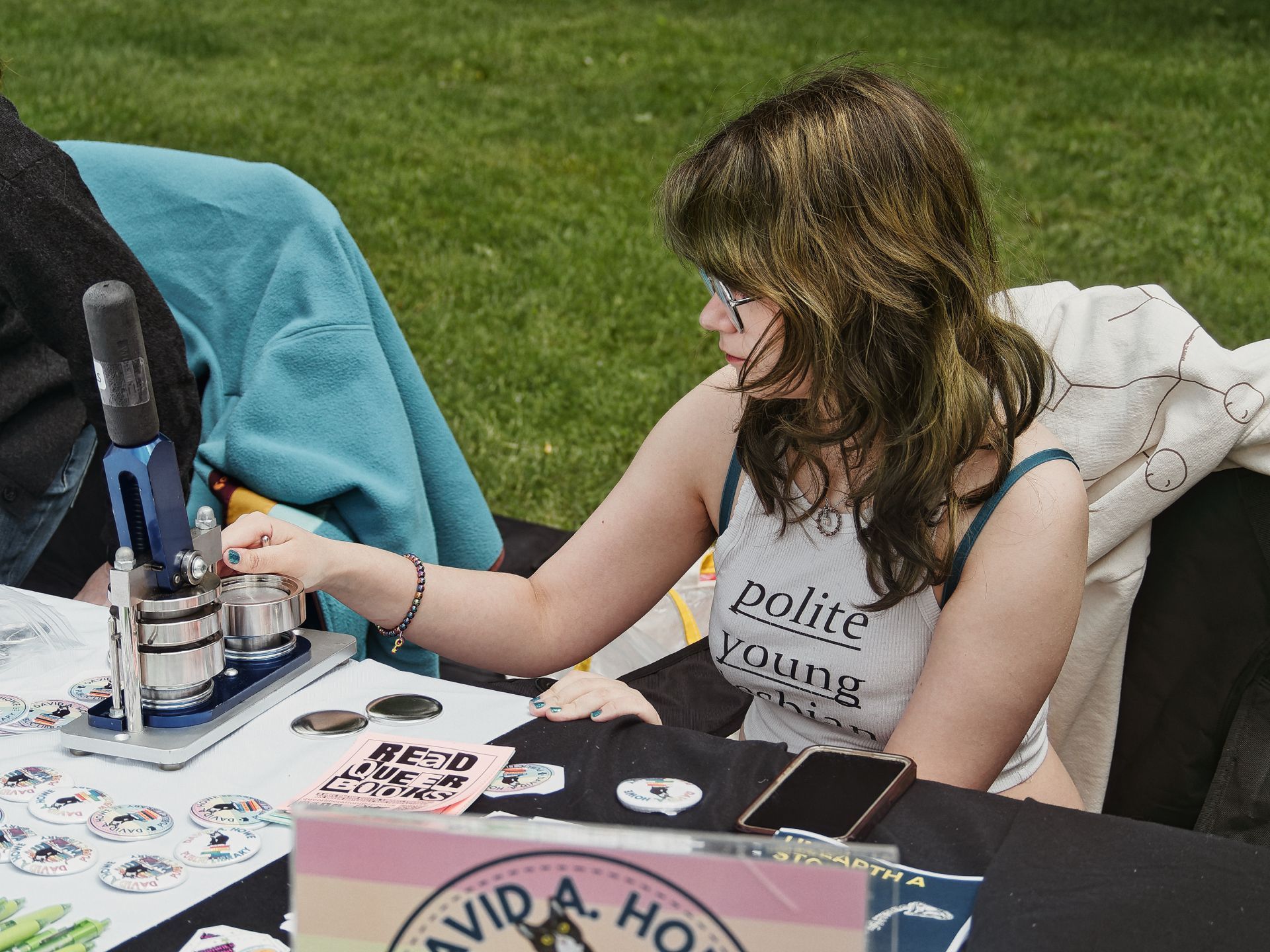 A young woman with wavy brown hair and glasses leans over an outdoor vendor table, operating a button-making press. Her tank top reads "polite young" with the rest of the text obscured. The table is covered in finished pinback buttons, a pink card reading "READ QUEER BOOKS," blank button blanks, and a partially visible sign reading "DAVID A. HO—"; green grass fills the background.