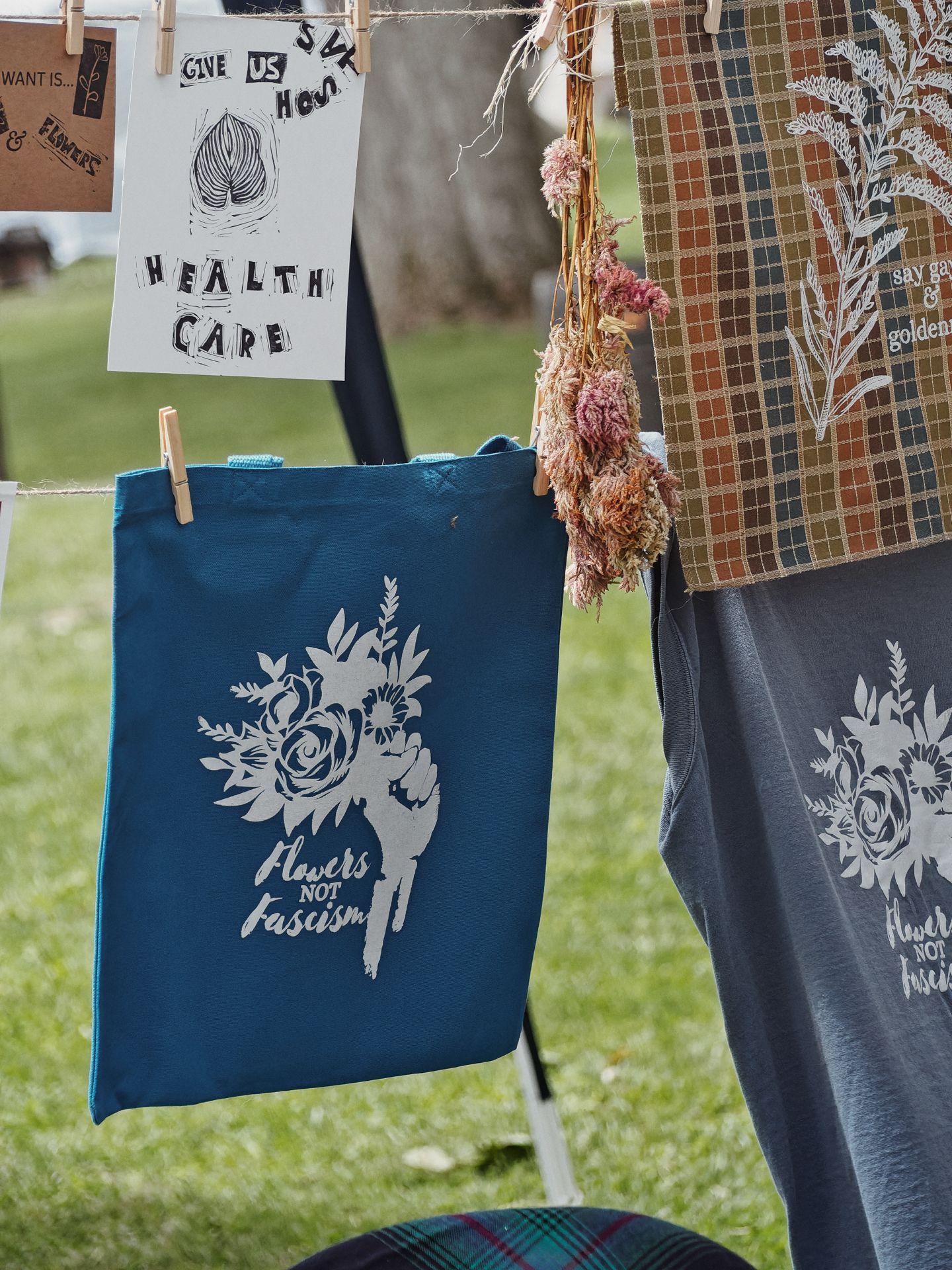 Outdoor market display with items clipped to a rack over a grassy background: a teal canvas tote printed with a white raised-fist bouquet design reading "flowers not fascism" hangs at center, beside a gray shirt with the same graphic; a handmade print clipped upper left reads "Give Us SVK How HEALTH CARE" with a leaf illustration; dried pink florals hang from twine at center, and a plaid earth-toned tote with a botanical print reading "say ga— & golden—" hangs to the right.