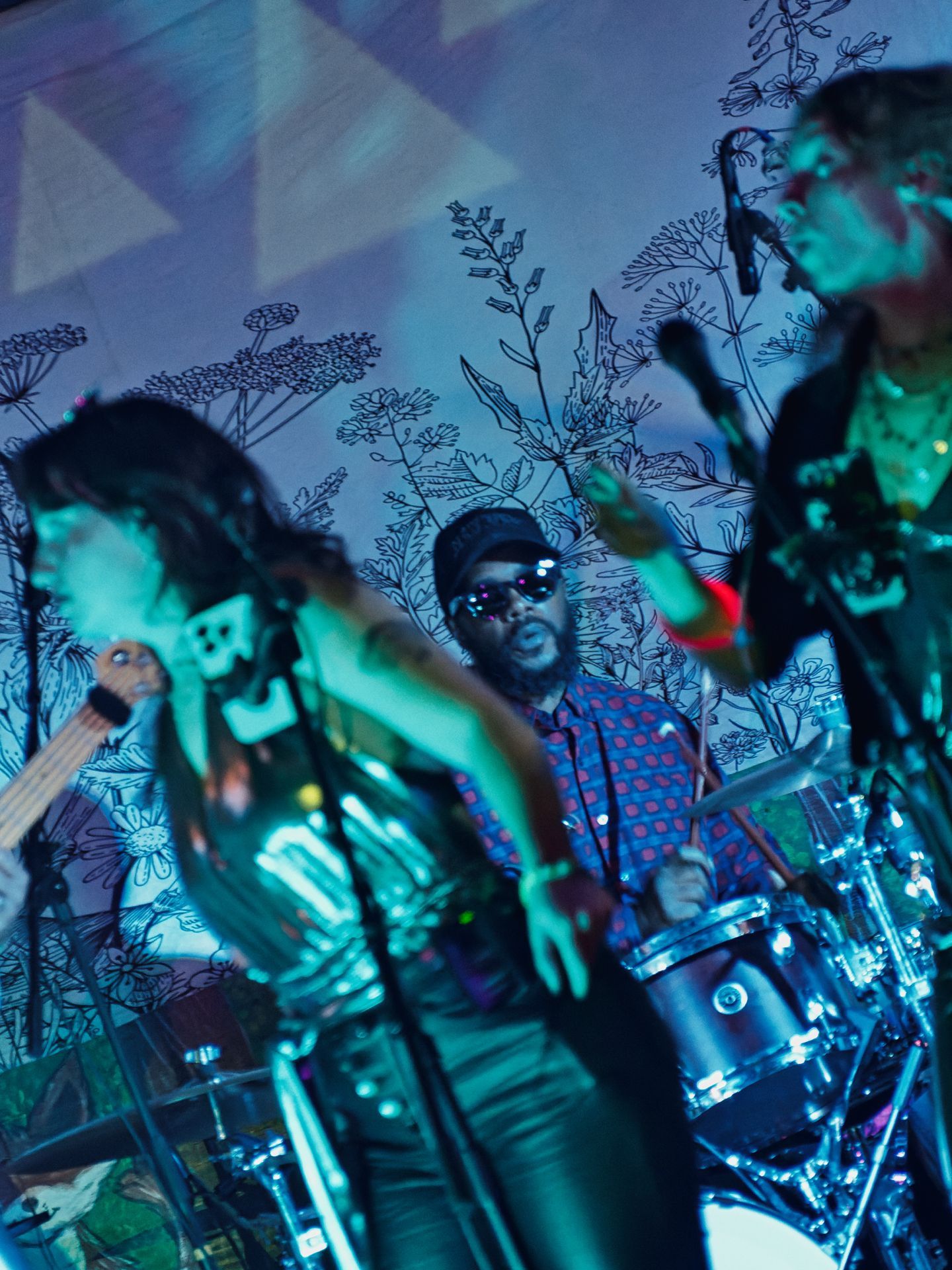 Vertical shot of a live band performing under deep teal and blue stage lighting, with a woman singing at a mic stand on the left, a man in mirrored sunglasses and a red-dotted blue shirt playing percussion in the center, and a second performer at the right edge. Behind them, a wall decorated with hand-drawn botanical line art and projected geometric triangles gives the venue an intimate, art-forward feel.