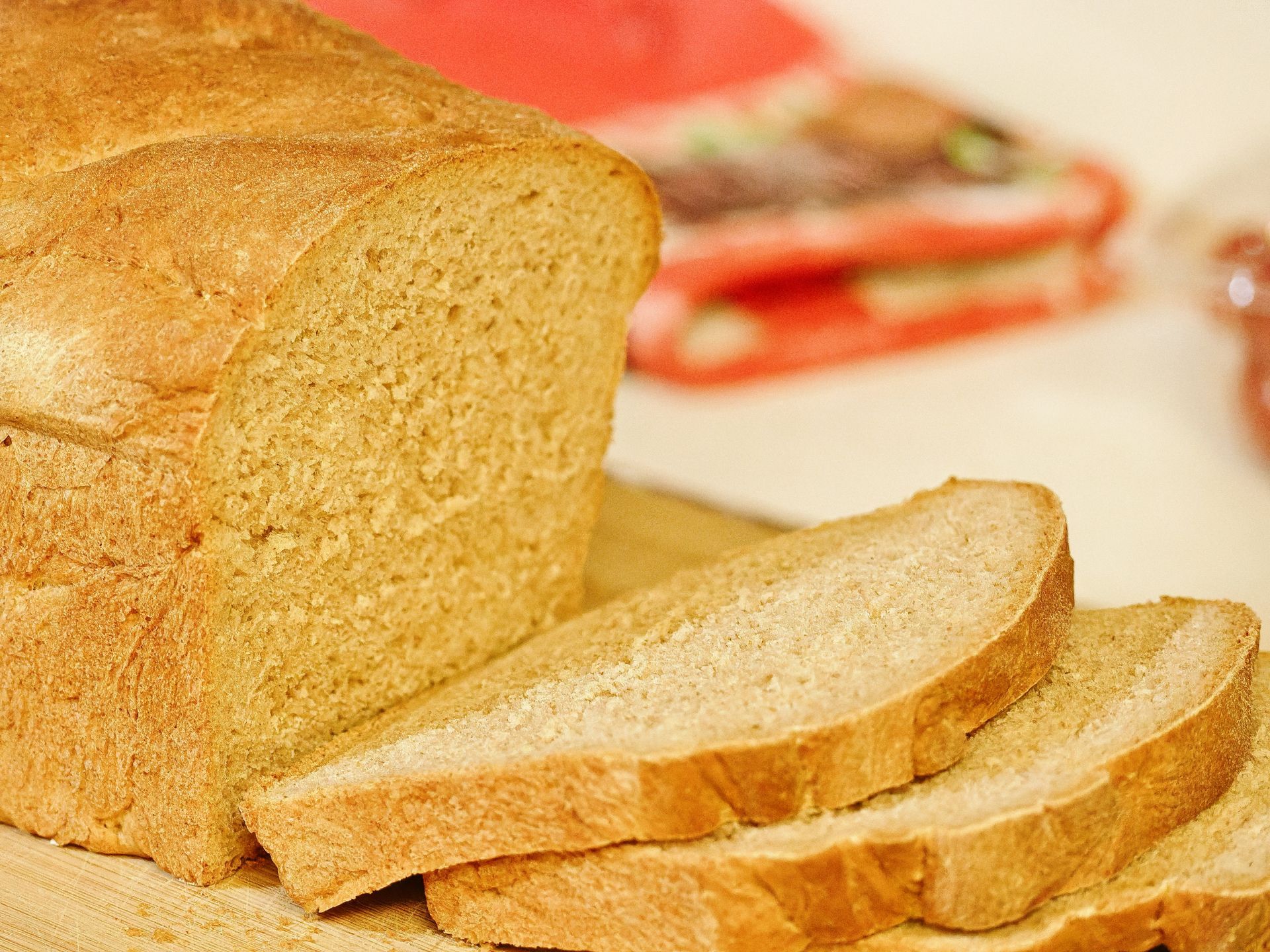 Golden-crusted whole wheat sandwich loaf on a wooden cutting board, with three slices fanned out to the right, revealing a fine, even crumb. A red-handled knife and indistinct items blur softly in the background.