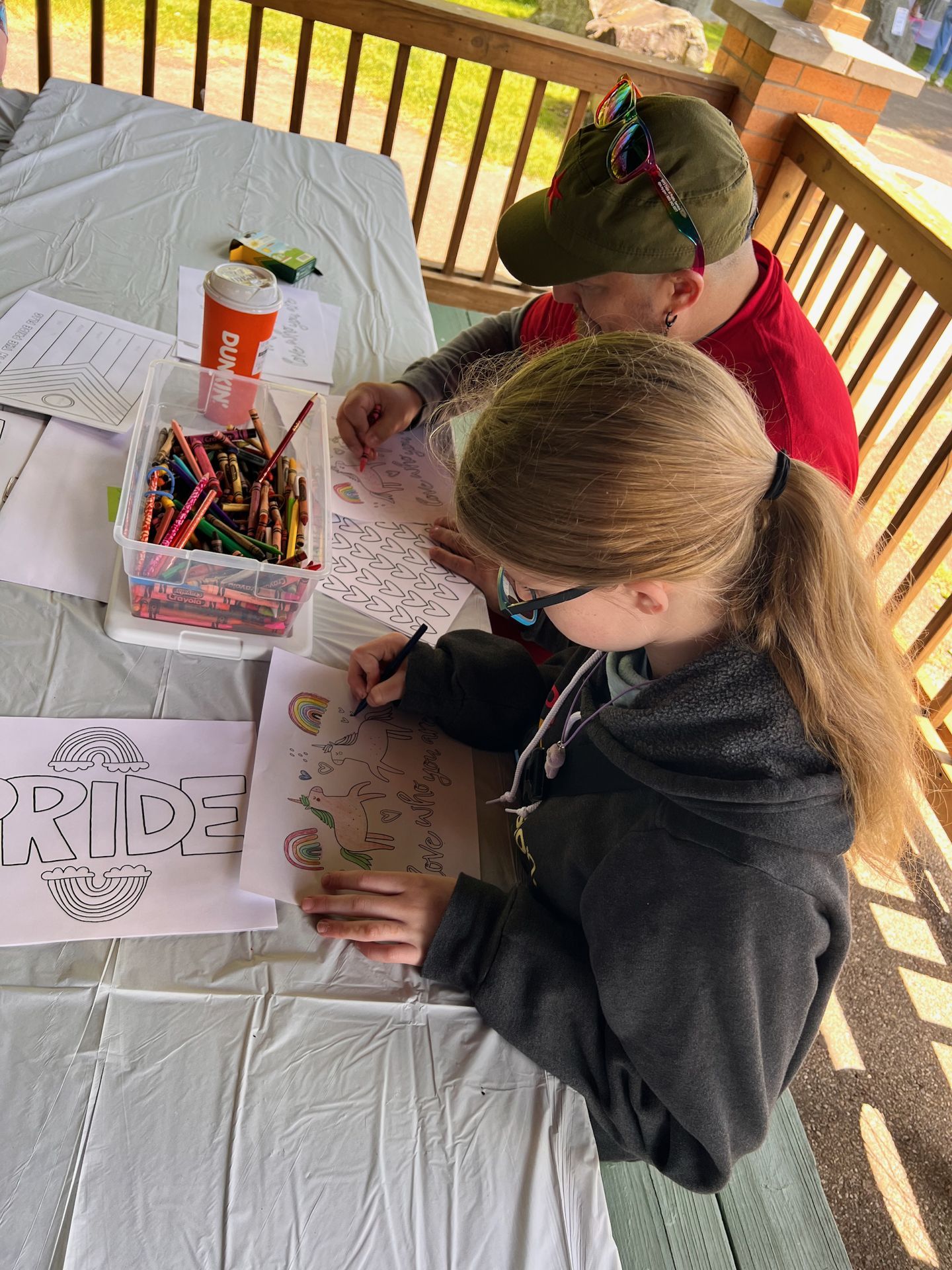 Two people coloring Pride-themed sheets at an outdoor table covered in a white tablecloth, with a clear bin overflowing with crayons and markers beside a Dunkin' cup. In the foreground, a young girl with long hair and glasses works on a sheet featuring a unicorn and rainbows; to her left, a partially visible coloring page reads "PRIDE" with a rainbow beneath it. Behind her, an adult in a red shirt and olive green cap with rainbow sunglasses colors another sheet covered in hearts.