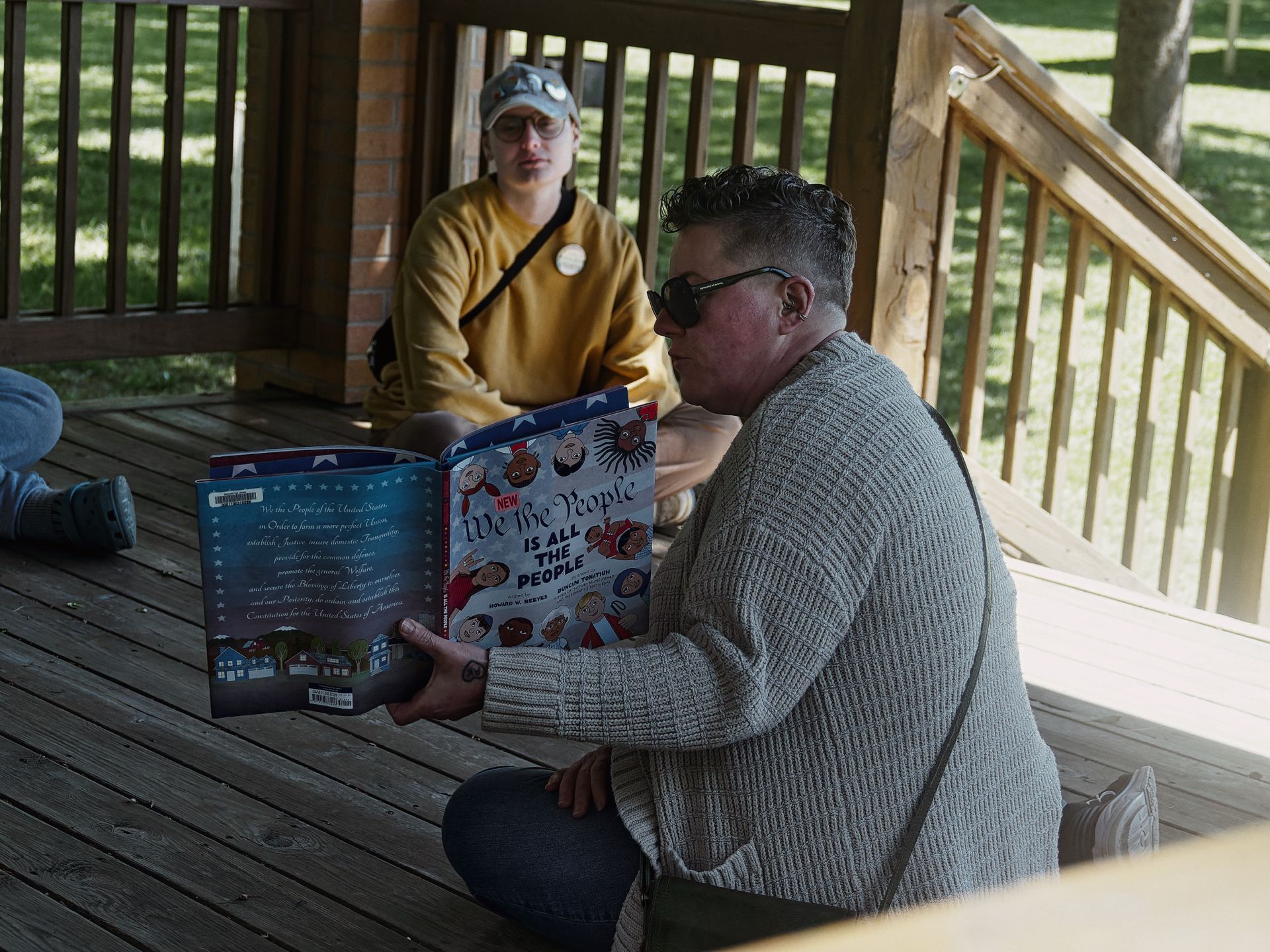 Person in a grey knit cardigan and sunglasses sitting on a wooden deck, holding open a children's book titled "NEW We the People IS ALL THE PEOPLE" — its illustrated cover showing diverse cartoon faces against a red, white, and blue background. A second person in a mustard yellow sweatshirt and grey cap sits cross-legged in the background, listening, with green grass visible beyond the deck railing.