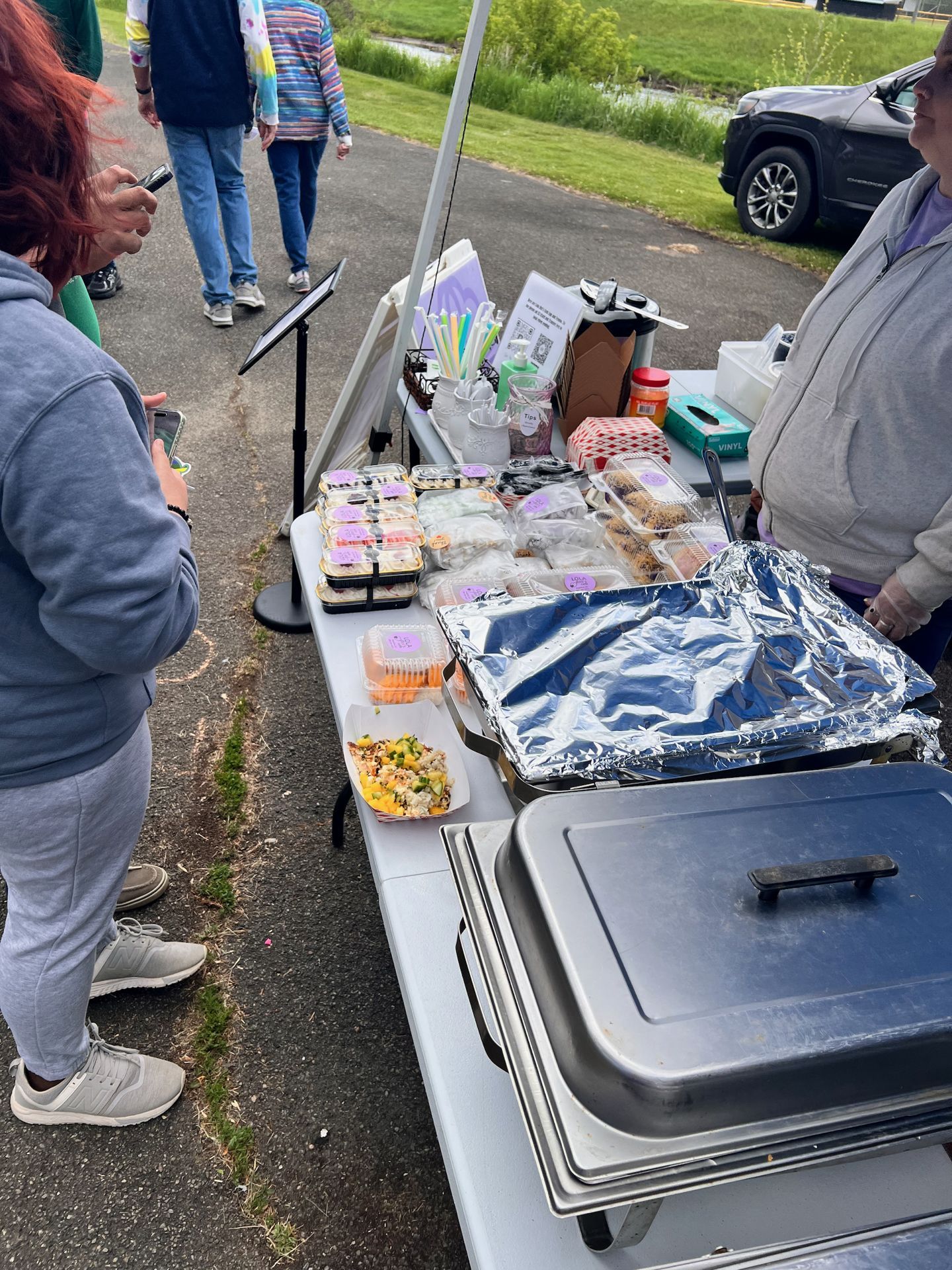 Vertical shot of an outdoor food vendor table under a canopy, lined with clear plastic clamshell containers of baked goods and prepared foods marked with small purple labels, alongside a foil-covered chafing tray and a dark chafing dish in the foreground. A red-haired person in a gray hoodie browses on the left, while a vendor in a gray hoodie stands on the right, with a parking lot and green grass visible in the background.